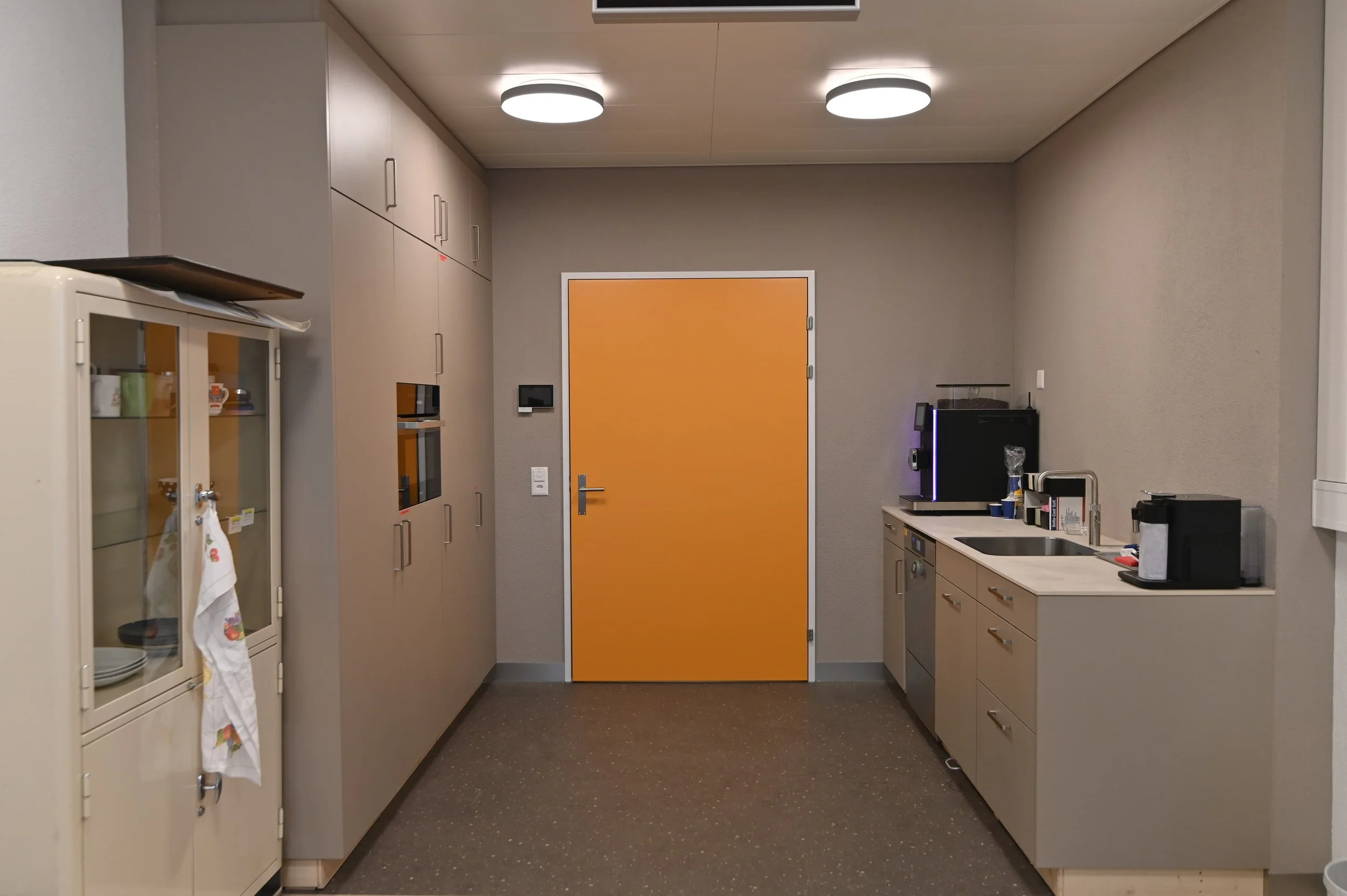 Büroküche mit orangefarbener Tür, weißen Schränken, Kaffeemaschine, Wasserkocher, Waschbecken und Kaffetasse auf der Theke, Deckenleuchten.