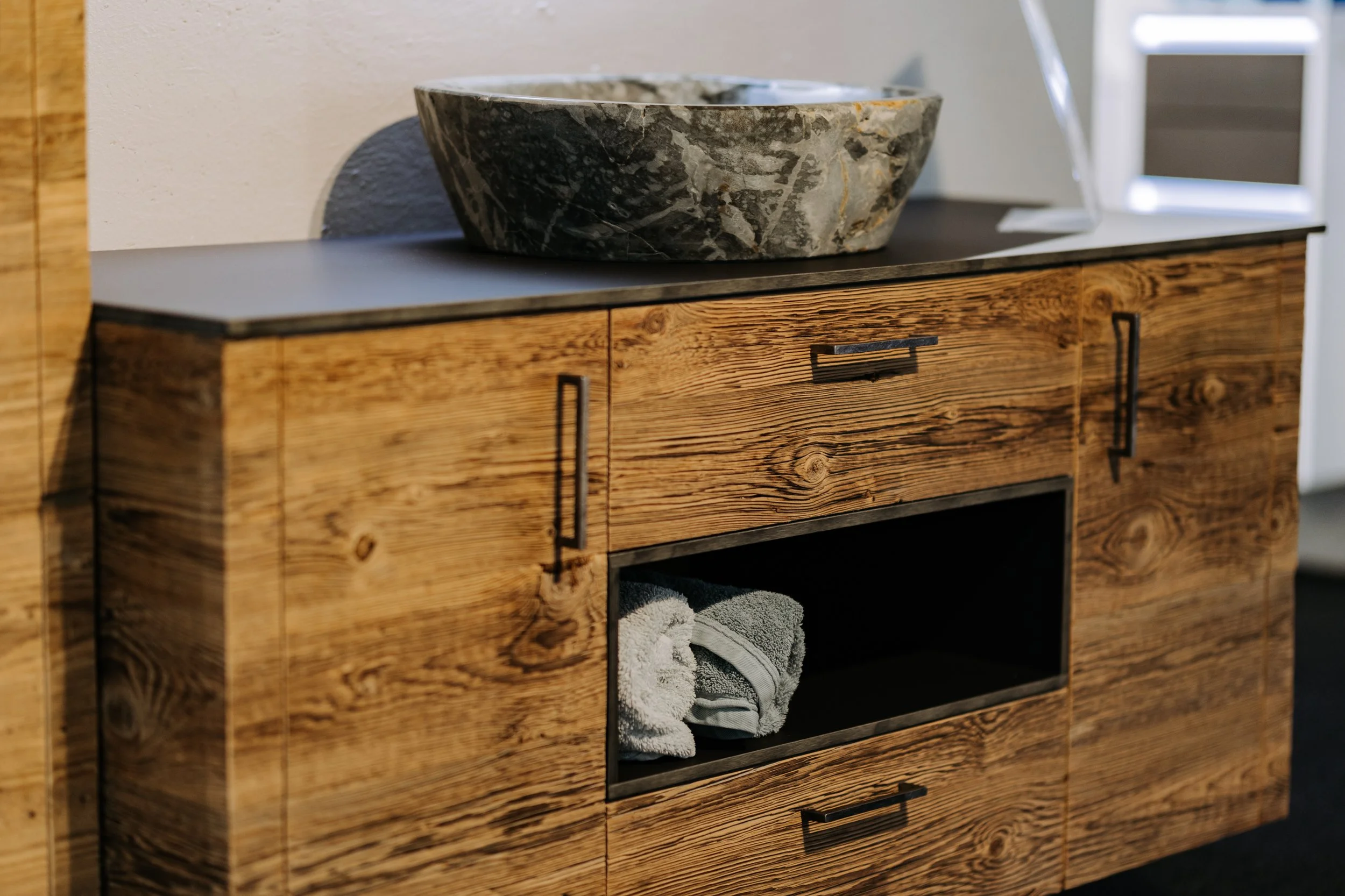 Holzschränkchen mit schwarzen Griffen und offenen Fächern, darin Handtücher, oben eine Schale aus dunklem Marmor auf heller Oberfläche.