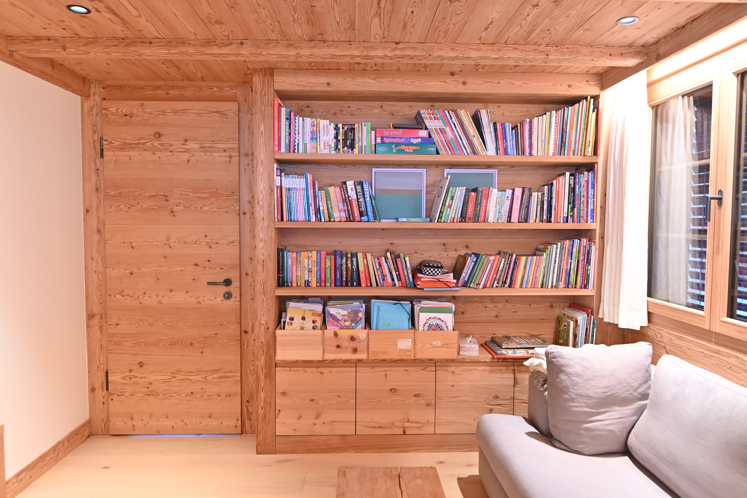 Ein Raum mit Holzwänden, einem großen Bücherregal voller Kinderbücher und einem Fenster mit Vorhängen. Im Vordergrund ein weißes Sofa.