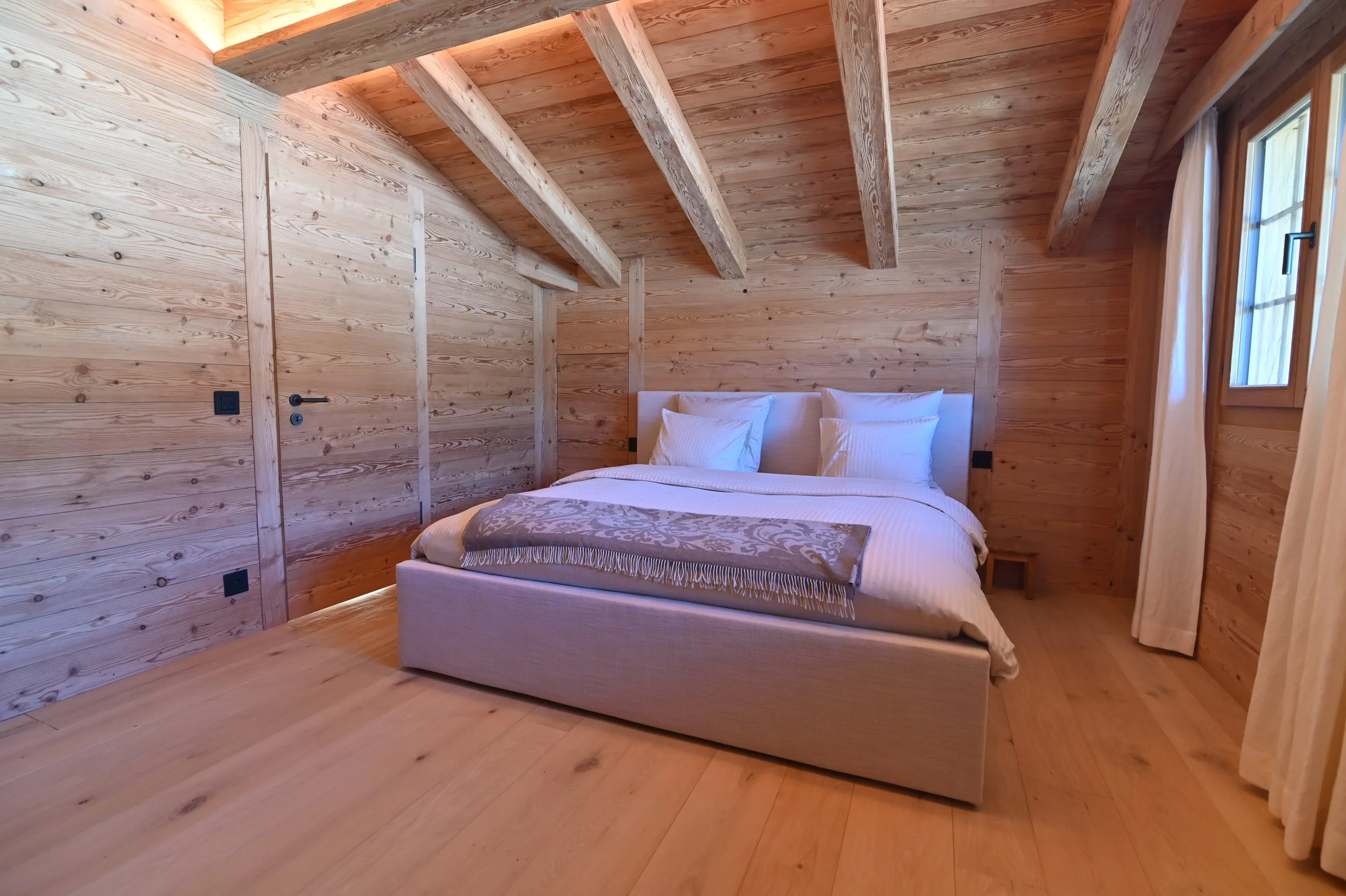 Ein Schlafzimmer mit Holzvertäfelung an Wänden und Decke, ein Doppelbett mit weißen Bettwäsche und Kissen, ein Fenster mit weißen Vorhängen.