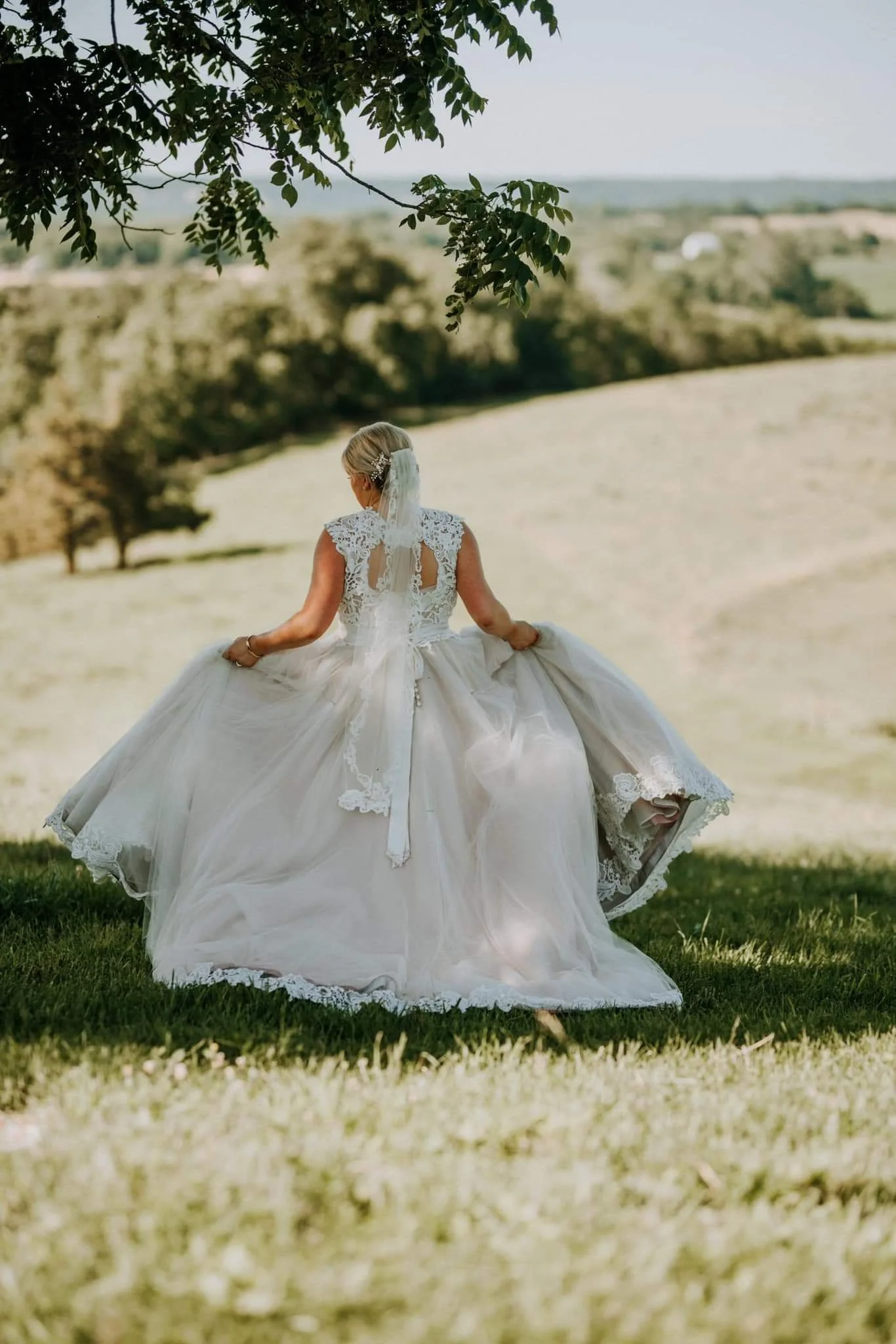 A bride in a white lace wedding dress outdoors, holding the sides of her dress, with a scenic landscape of trees and rolling hills in the background.