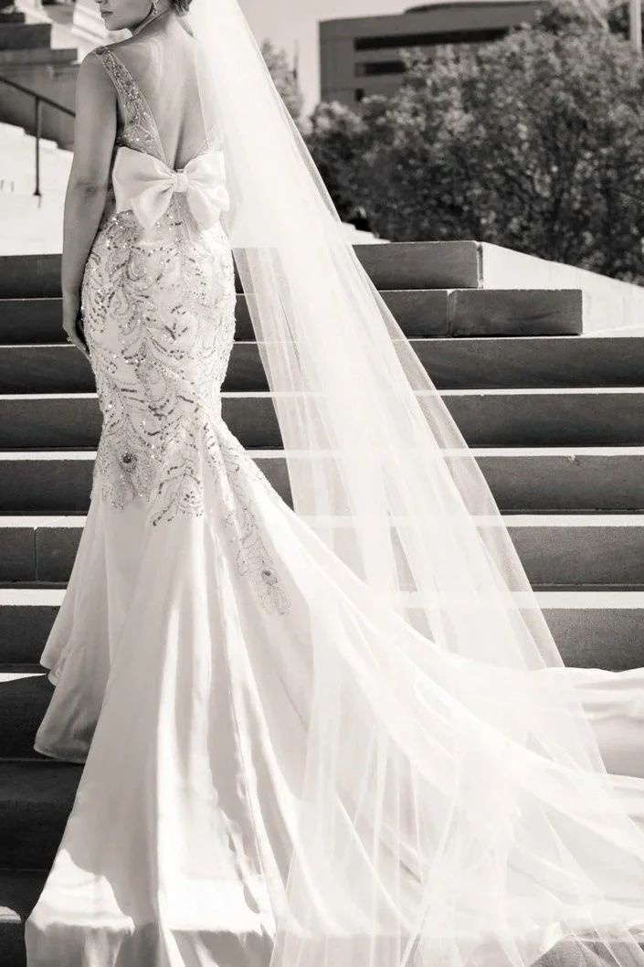 A bride in a detailed wedding gown with a long train and a bow on the back, standing on outdoor stairs with trees and buildings in the background.