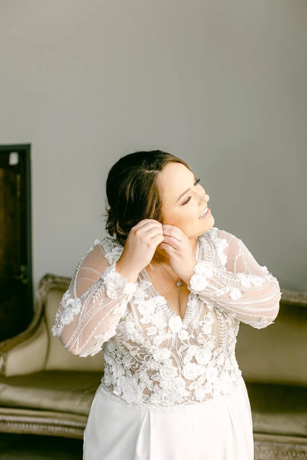 A woman in a white lace wedding dress putting on earrings, smiling and looking to the side while standing in a room with a vintage sofa in the background.
