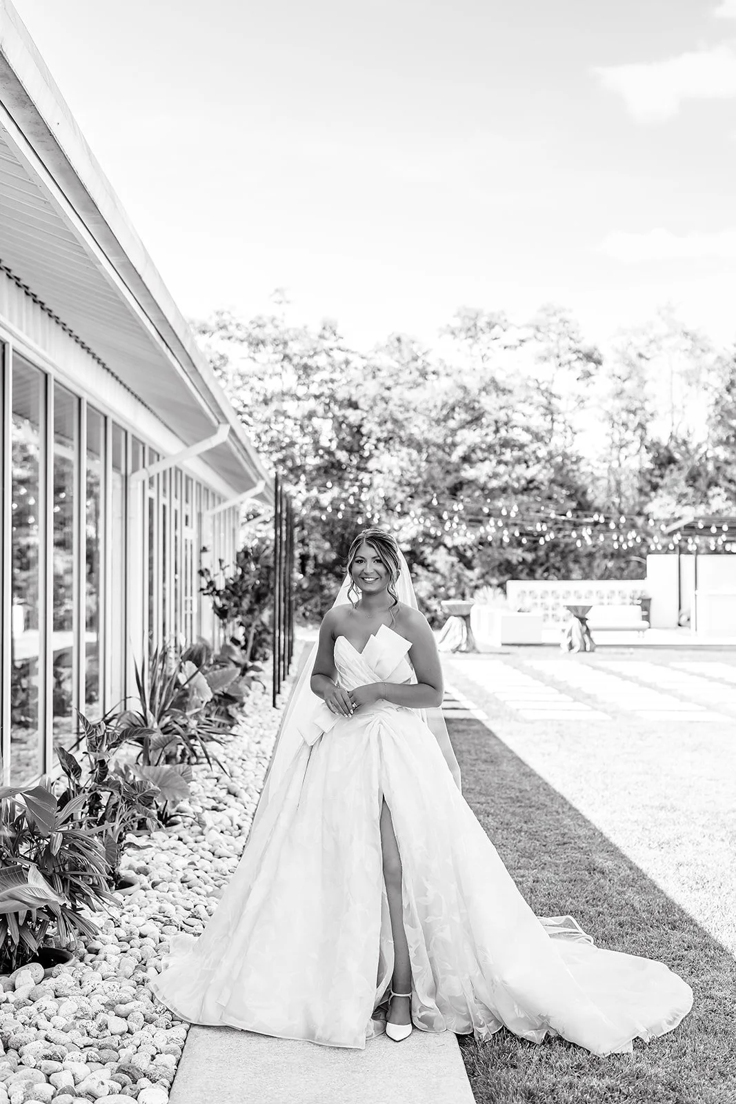 A woman in a wedding dress stands outdoors, smiling, next to a modern building with glass walls, with trees and string lights in the background.