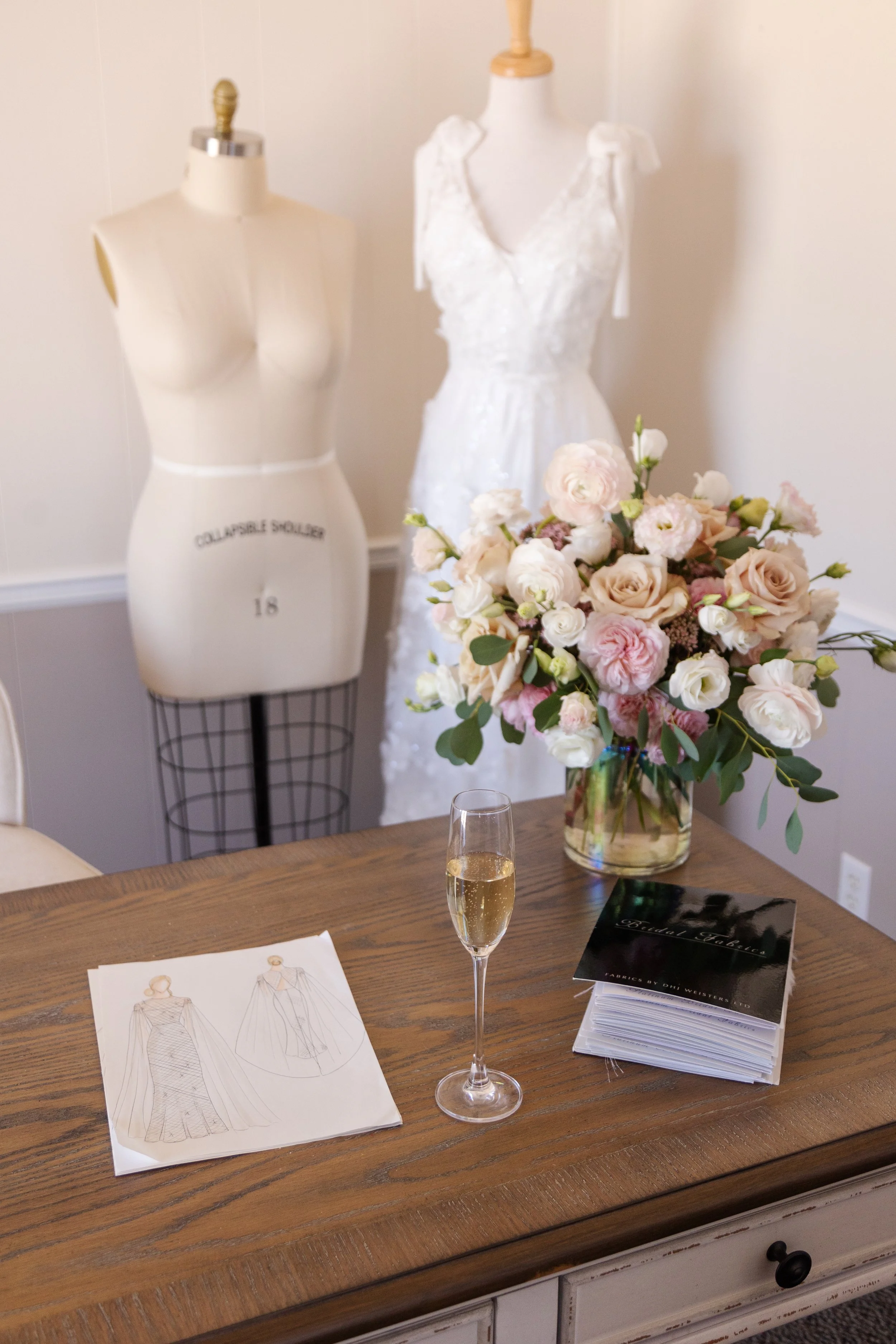 A table with a bouquet of pink and white flowers, a glass of champagne, a fashion sketch, and a booklet, with a wedding dress and mannequin in the background.