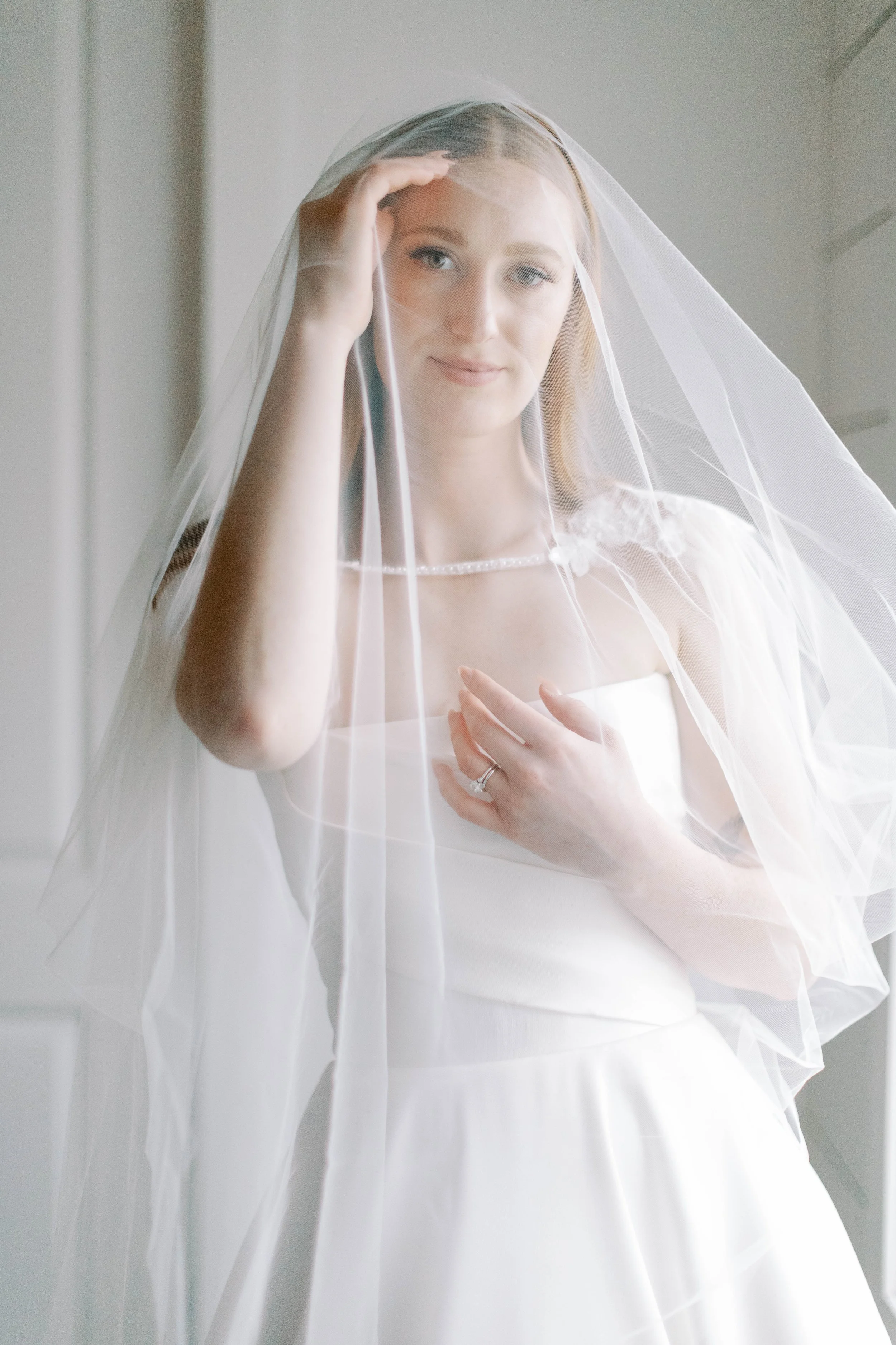 A bride in a white wedding dress and veil, standing indoors, looking at the camera, with her hand on her chest.