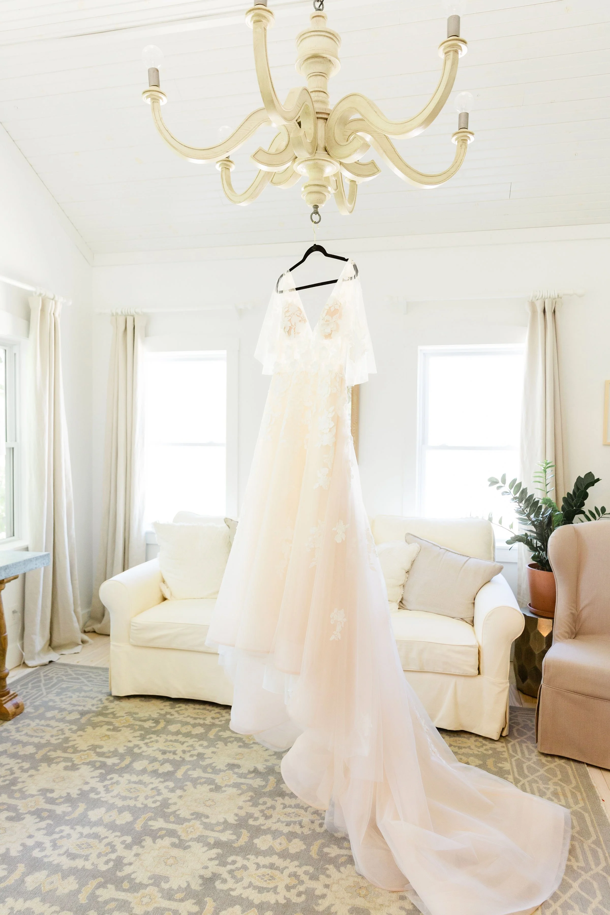 A wedding dress hanging from a black hanger on a chandelier in a bright, white living room with beige curtains, a white sofa, and a potted plant.
