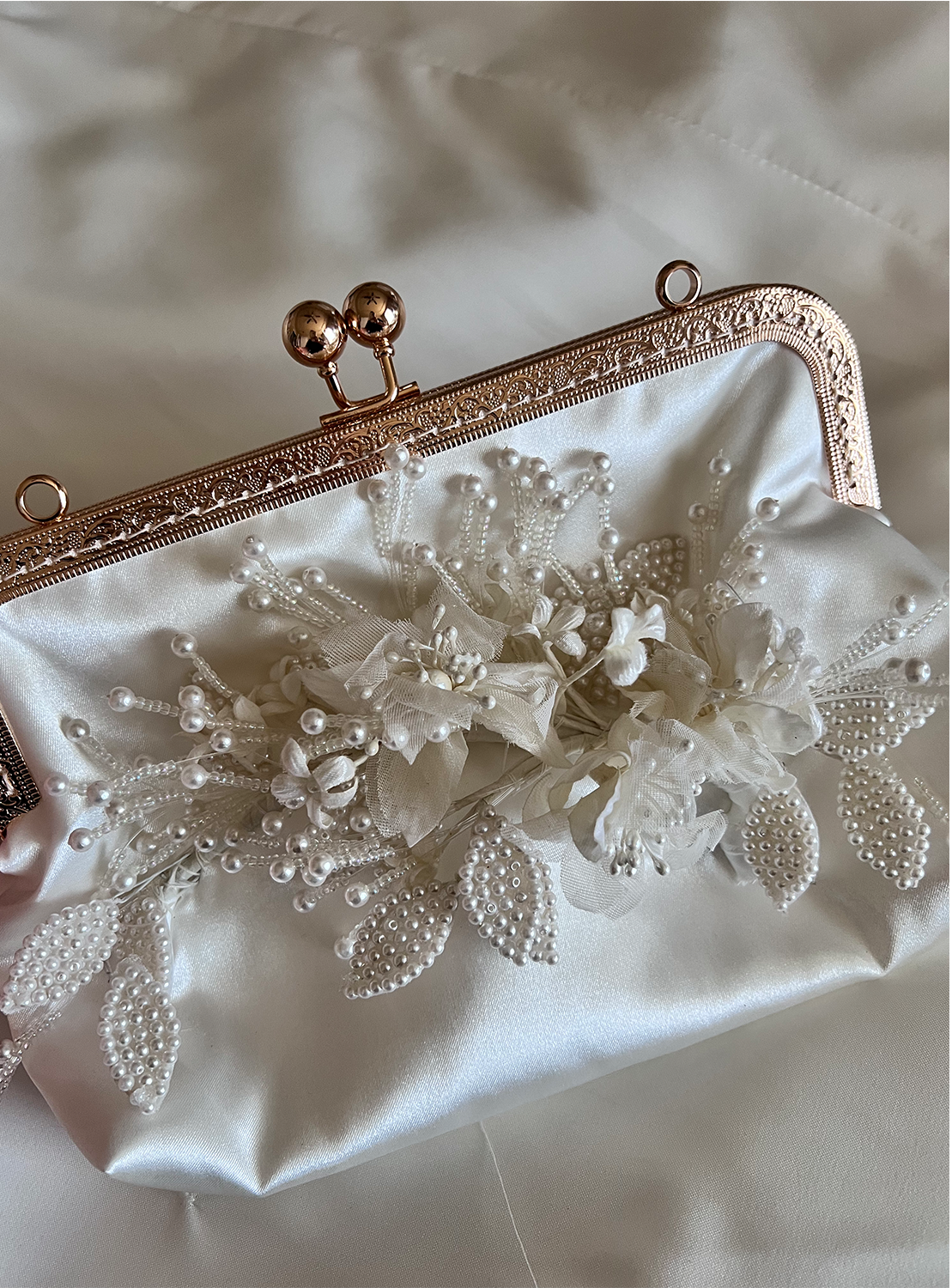 Cream-colored satin clutch bag with a decorative pearl and fabric floral design inside, featuring a rose gold metal frame with a ball clasp.