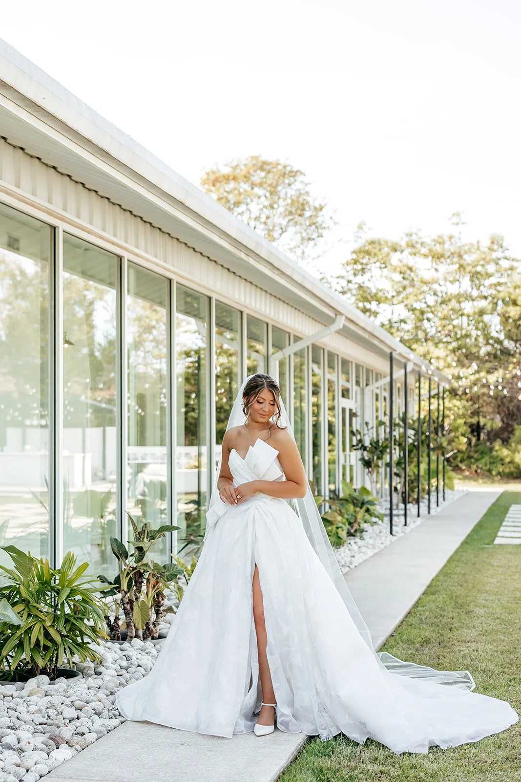 Bride in a white wedding gown standing outside near a glass wall with greenery.
