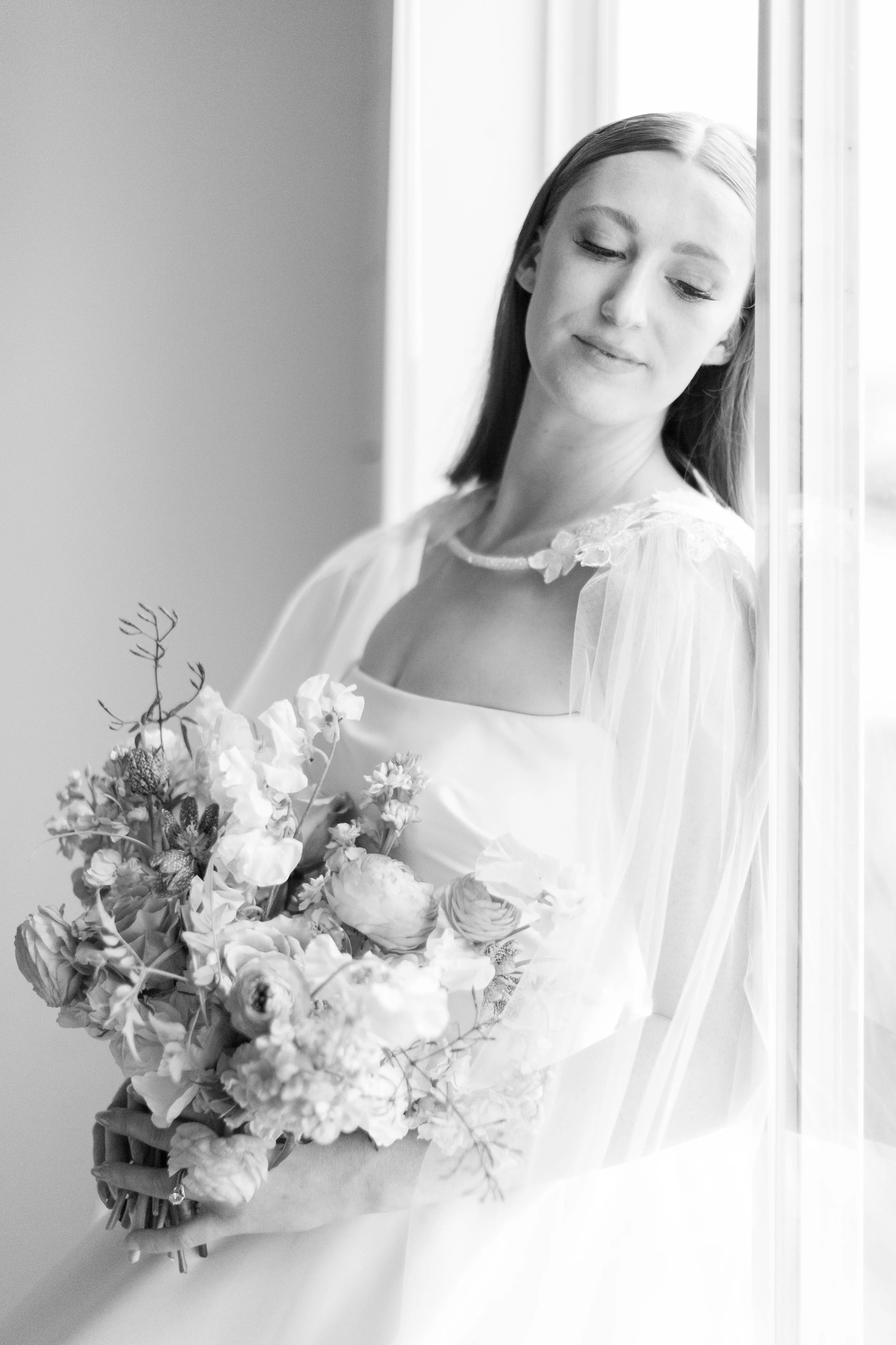 Black and white photo of a bride holding a bouquet of flowers, with a gentle smile, leaning against a window.