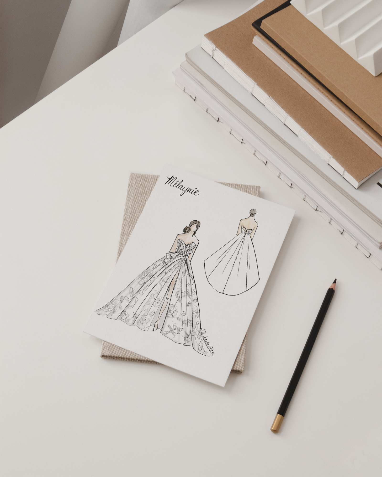 Design sketches of a wedding dress on white paper, placed on a beige notebook, with a black pencil nearby, on a white desk with stacked notebooks and a decorative white vase in the background.
