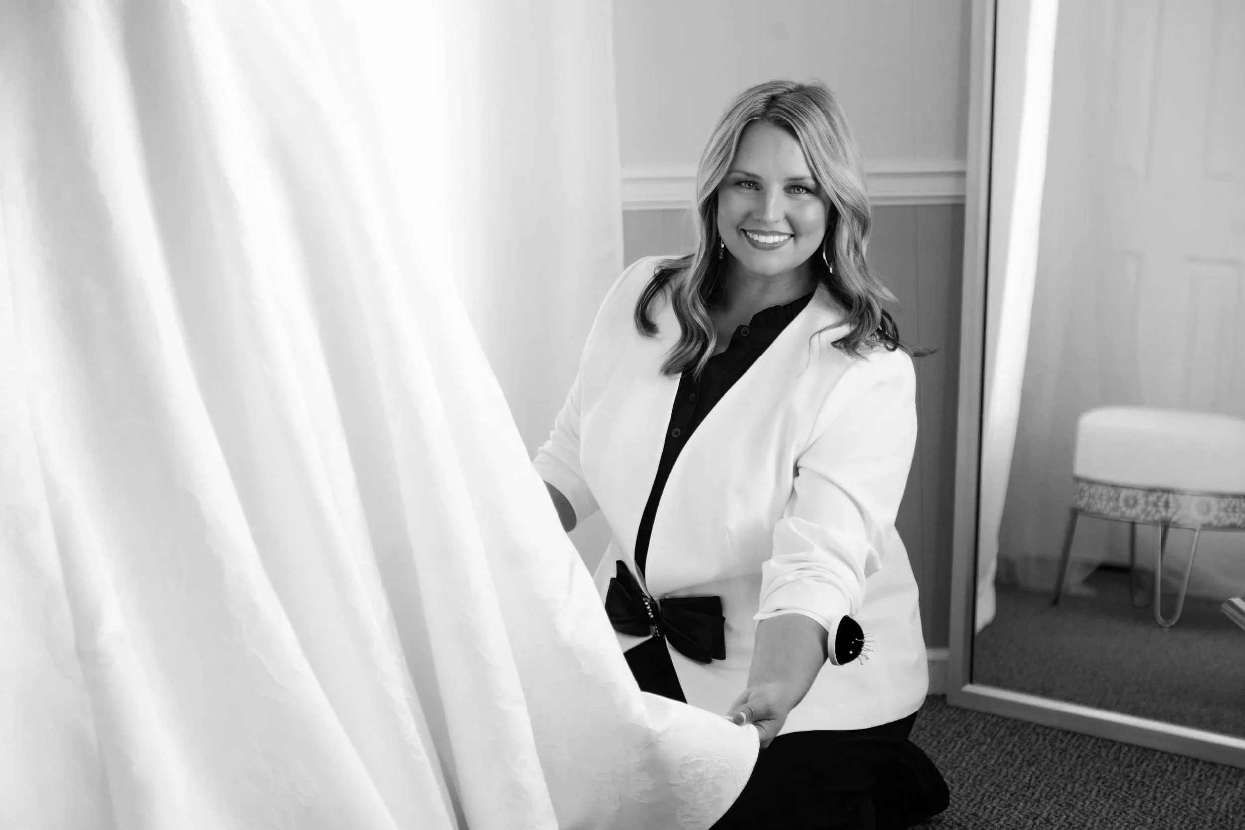 A woman smiling while holding a wedding dress in a fitting room.