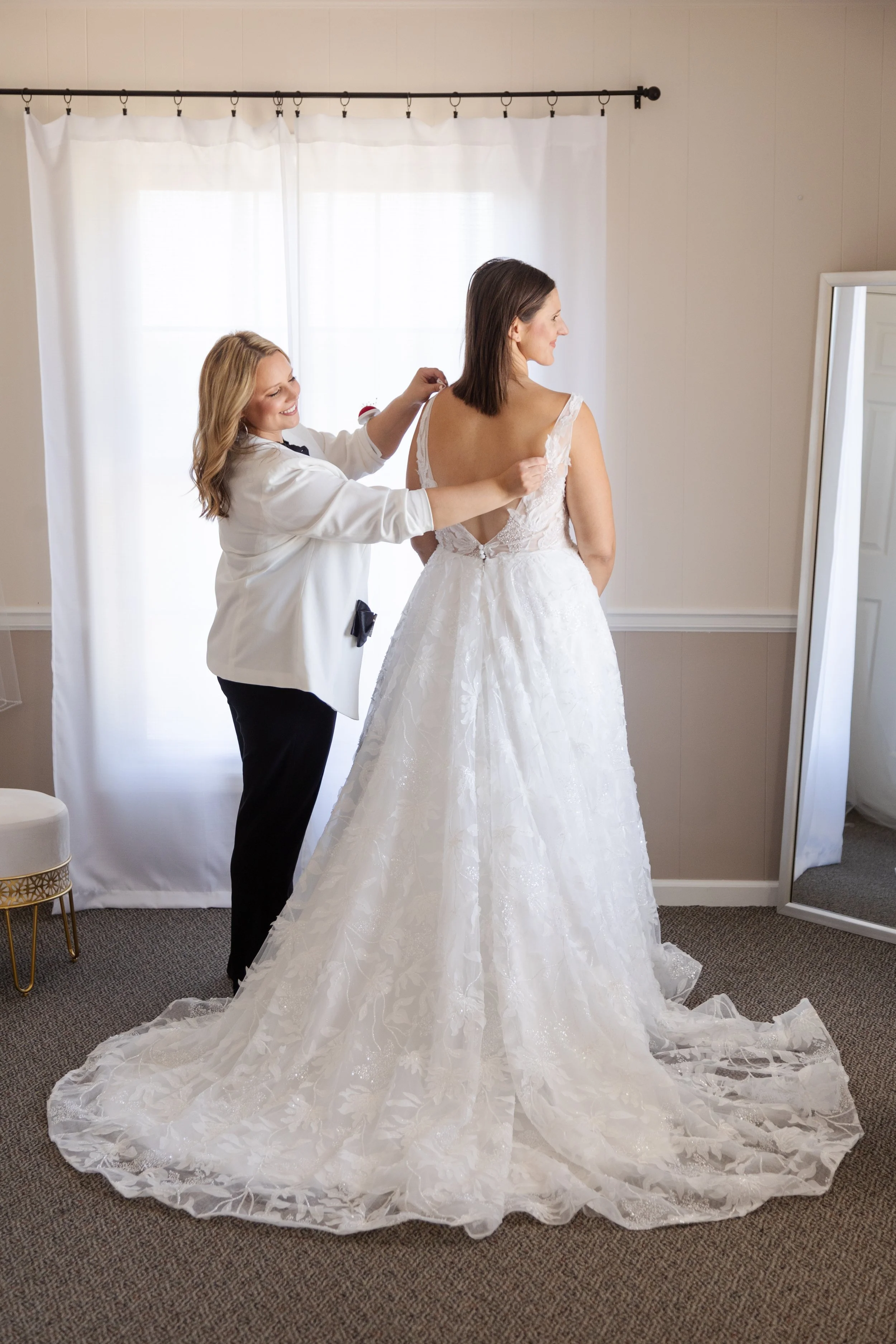 A bride in a wedding dress getting ready, with a woman helping her from behind, in a room with white curtains and a full-length mirror.