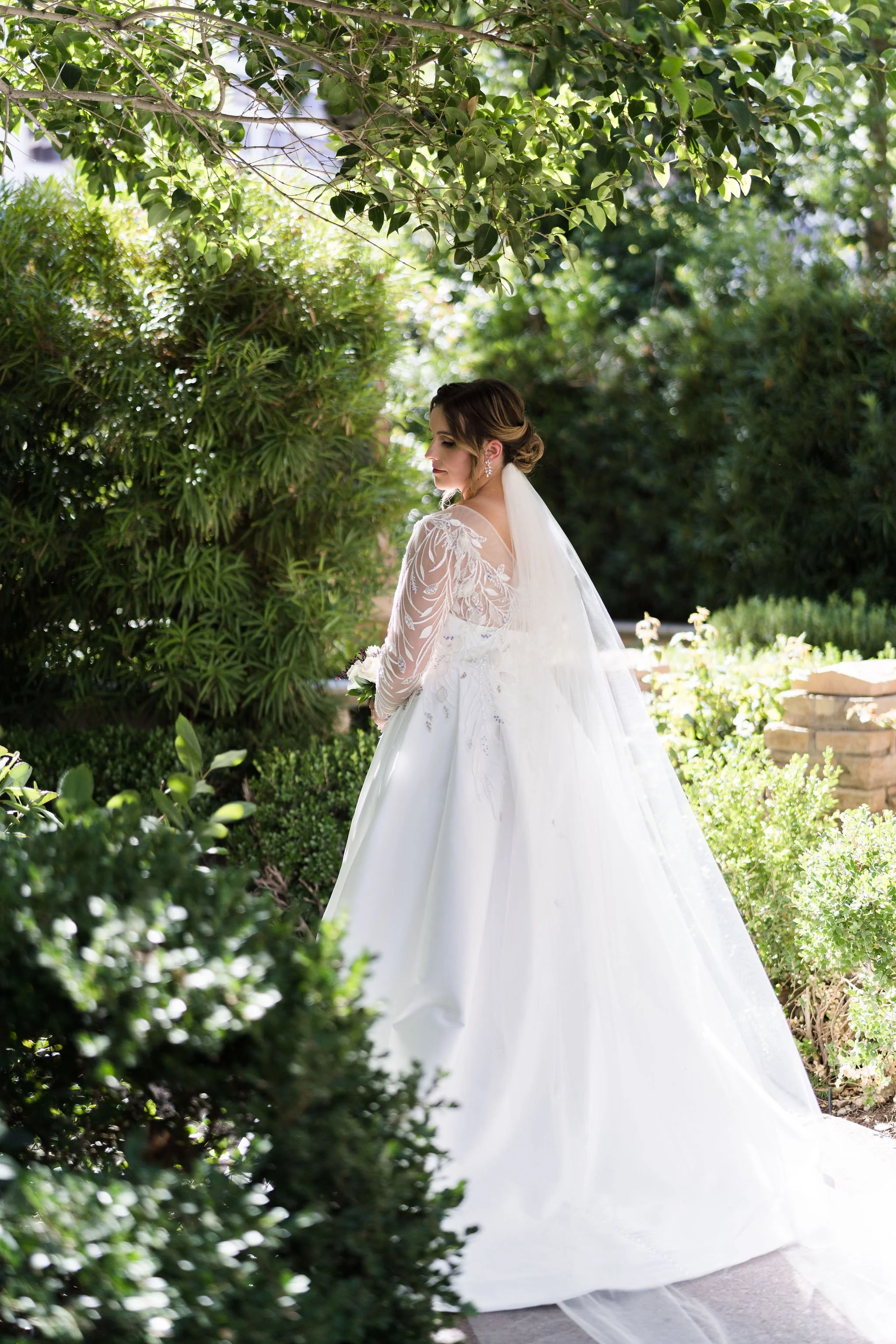 A bride in a white wedding dress standing outdoors in a garden with green foliage and plants.