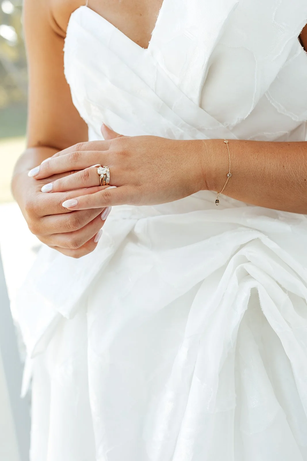 A woman in a white dress with her hands clasped, wearing a diamond ring on her ring finger and a delicate gold bracelet on her wrist.