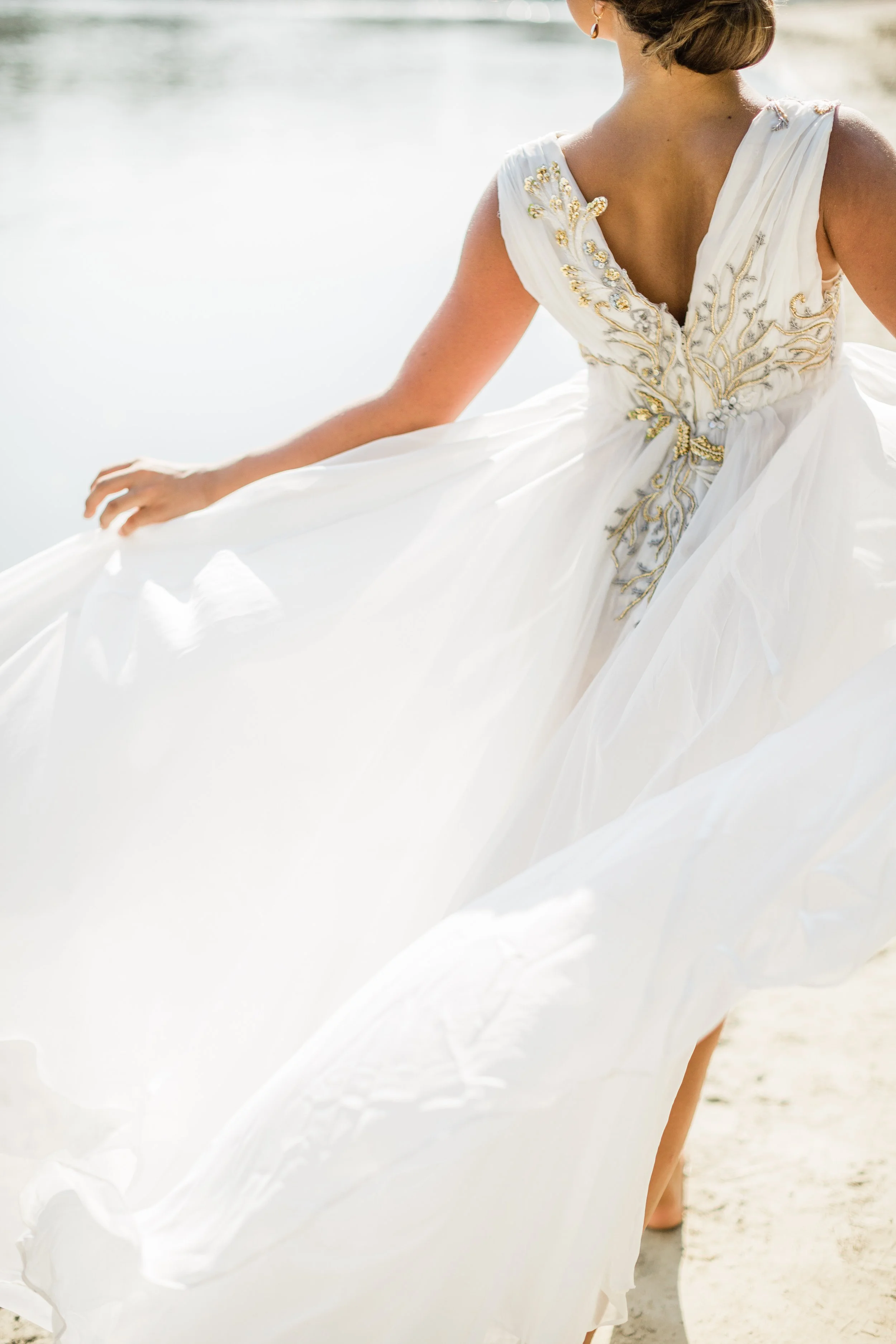 A woman in a white wedding dress with gold embellishments standing on a sandy beach by the water.