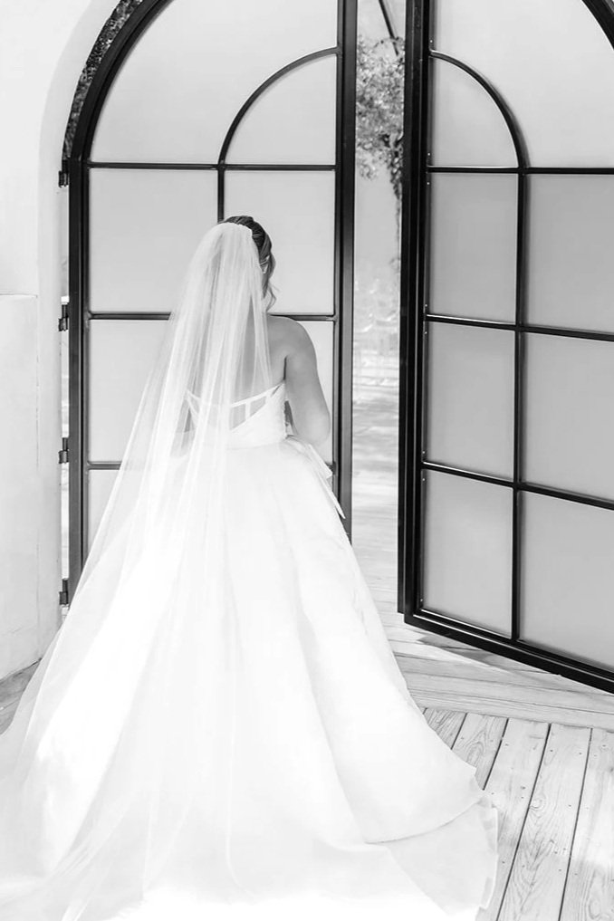 A bride in a wedding dress and veil looking out through large glass doors.