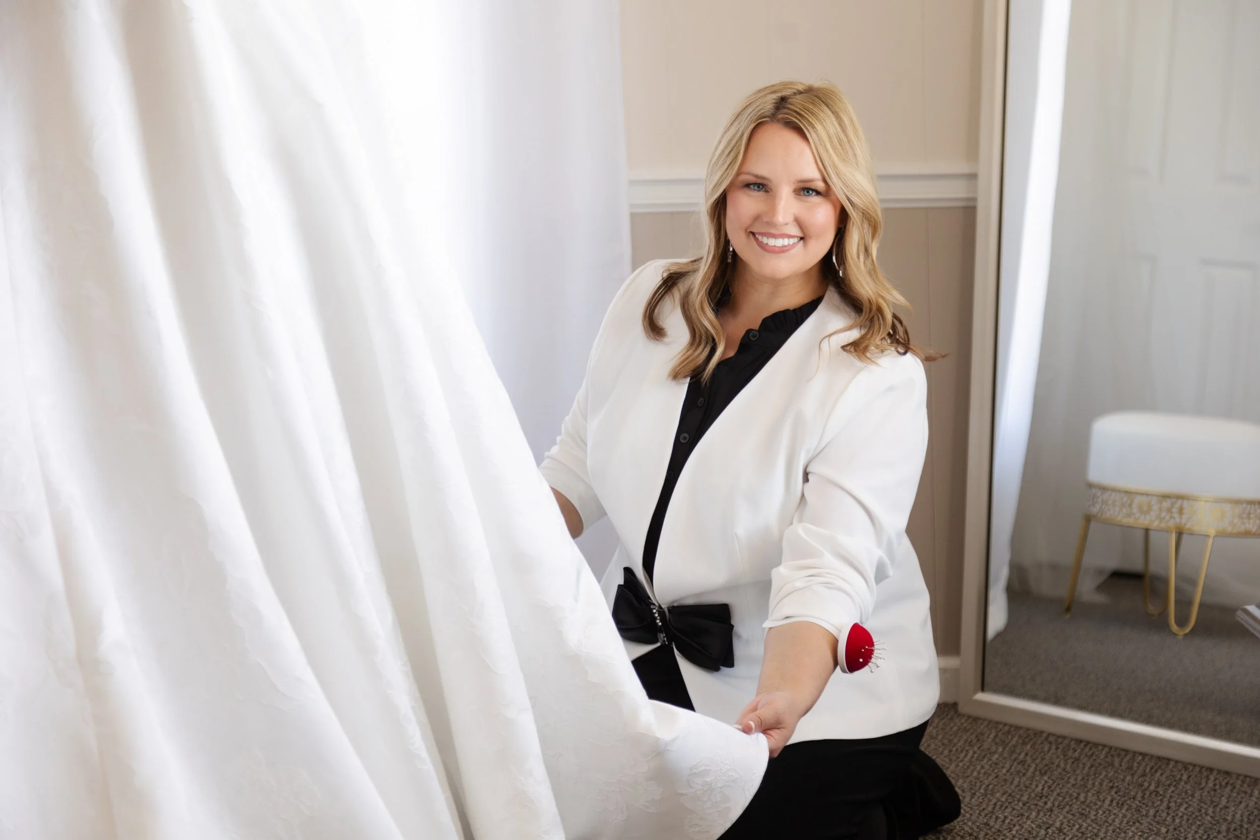 A woman with blonde hair smiling at the camera while holding a white curtain in a fitting room.