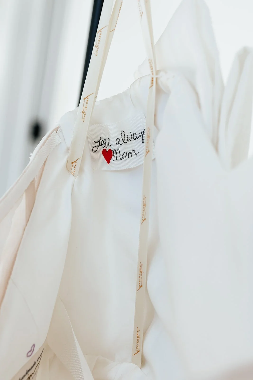 White fabric with a sewn-in label that reads 'Love always Mom' with a red heart, hanging on a clothing rack with beige ribbons.