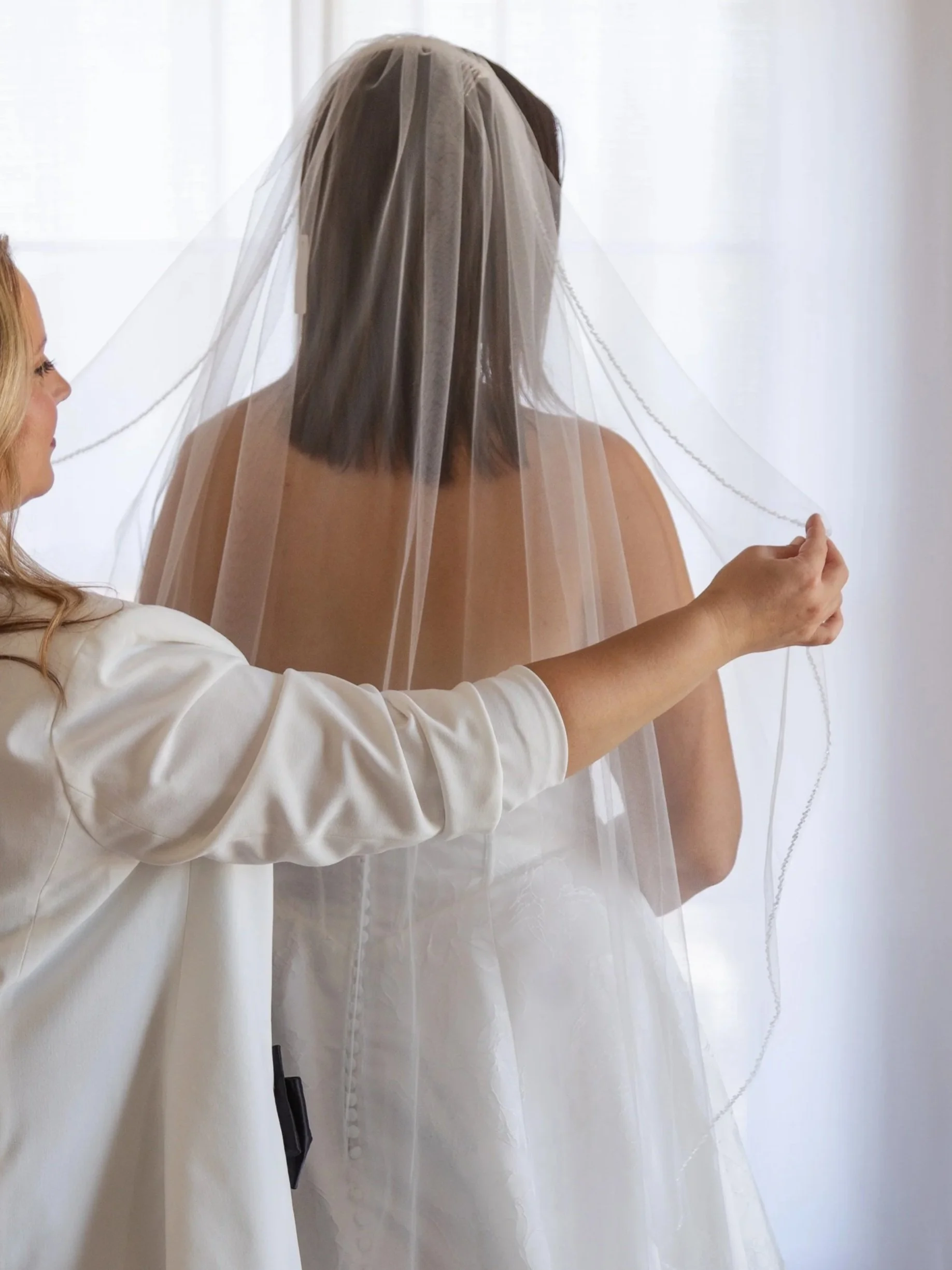 A woman in a white wedding dress with a veil being adjusted by another woman.