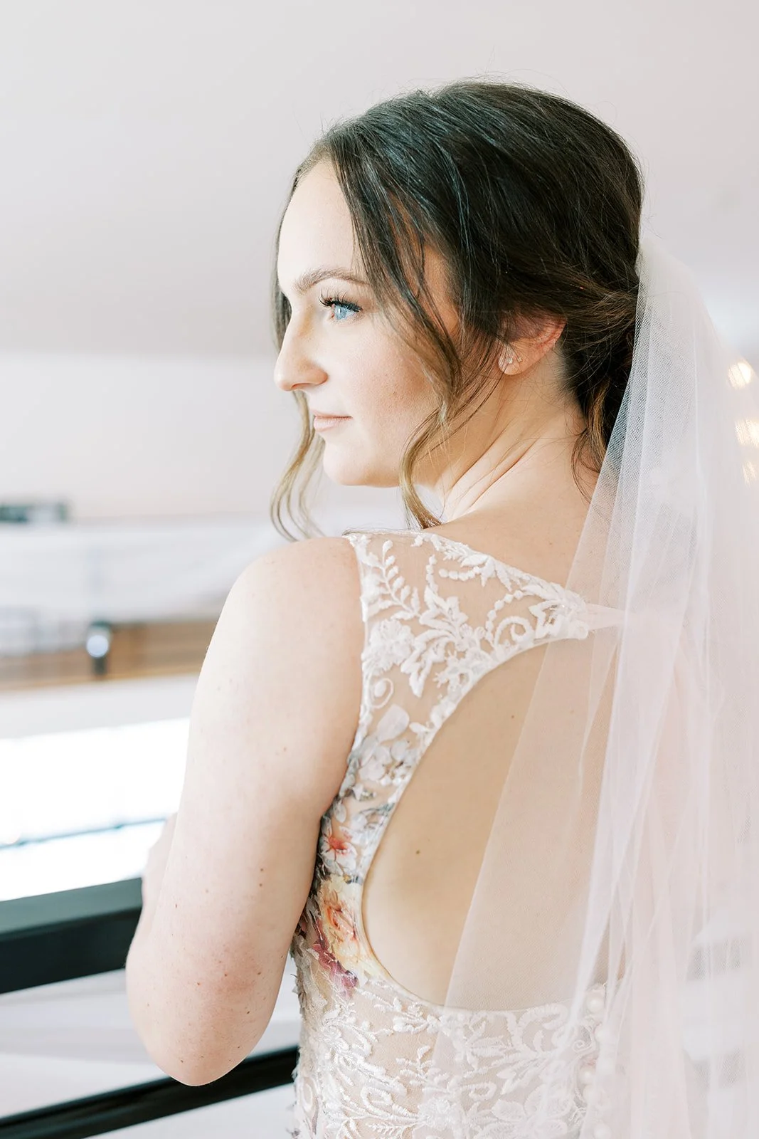 Close-up of a bride in a lace wedding dress with floral embroidery, looking to the side, with veil and a soft background.