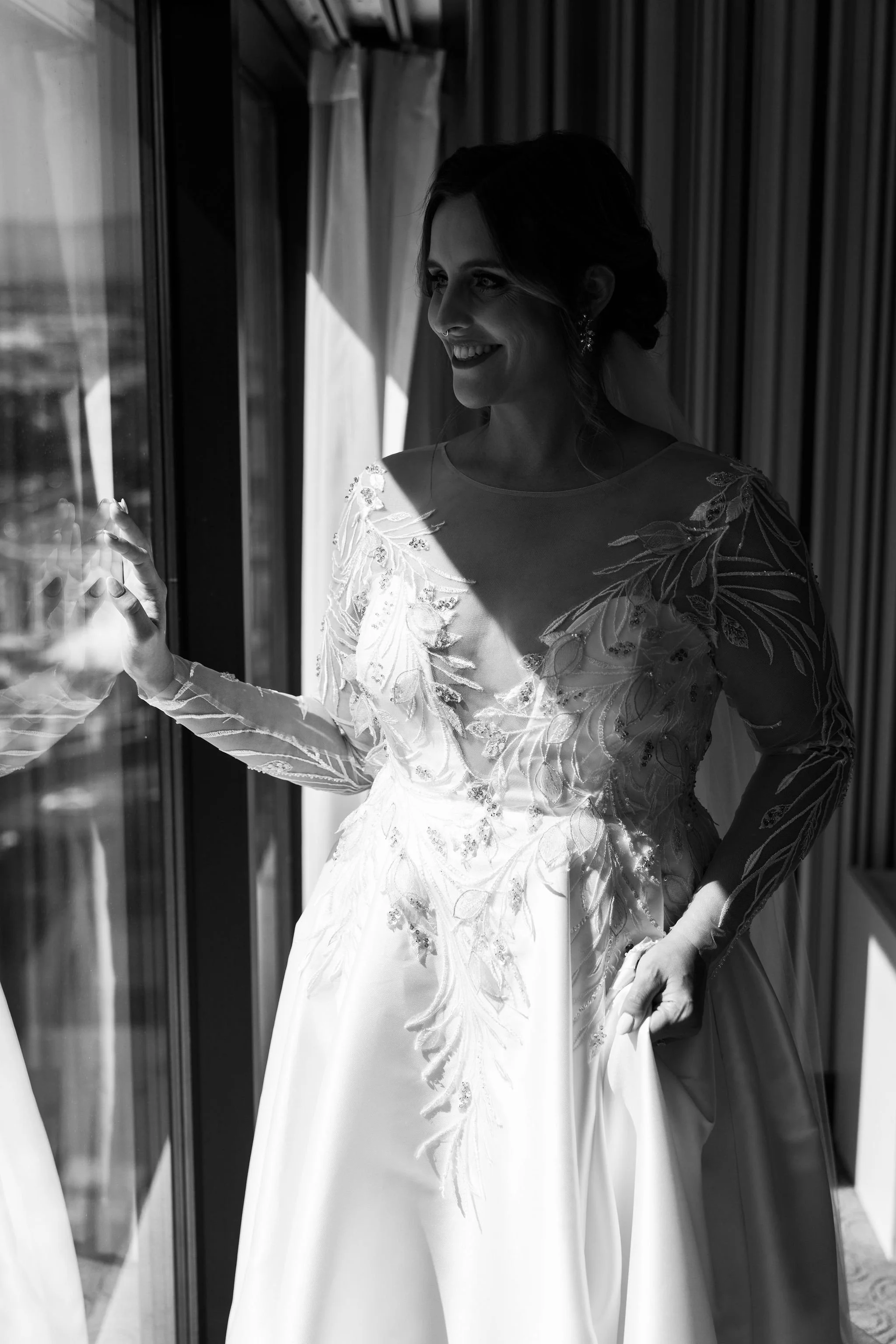 A smiling woman in an elegant wedding dress with embroidery, standing by a window with her hand on the glass, and sunlight illuminating her face.