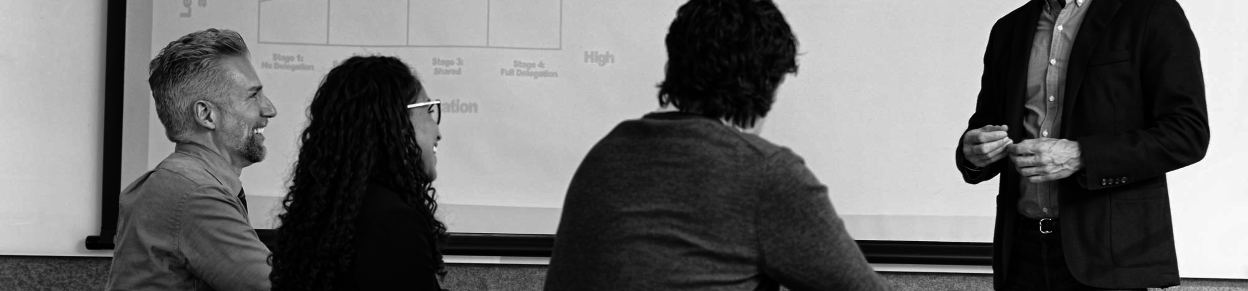 A man in a suit is standing in front of three seated people, giving a presentation to a group in a conference room.