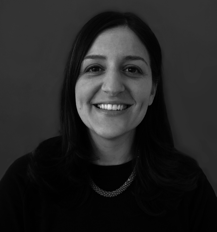 Black and white portrait of a woman with long dark hair, smiling, wearing a dark top and a pearl necklace, against a plain background.
