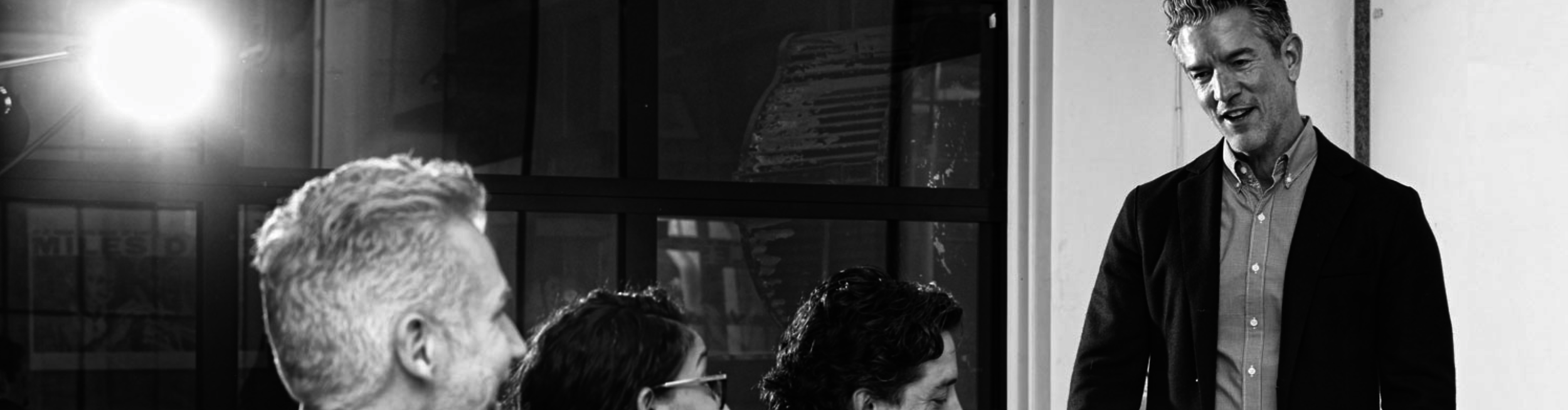 A man in a suit is standing and speaking to a group of seated people in an office setting. The group consists of adults, some wearing glasses, listening attentively. The background shows large windows and some office furniture.