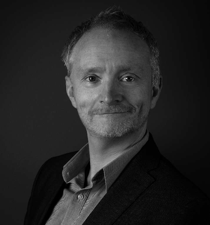 A grayscale portrait of a middle-aged man with short, light-colored hair and a beard, wearing a suit jacket and a collared shirt, smiling slightly against a plain background.