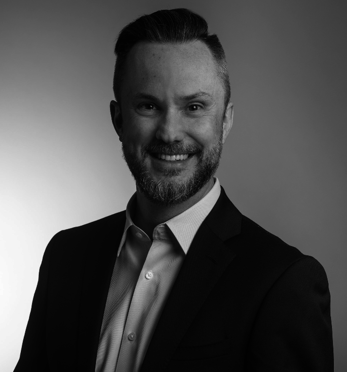 Black and white headshot of a smiling man with neatly styled hair and a beard, wearing a suit jacket and a collared shirt.