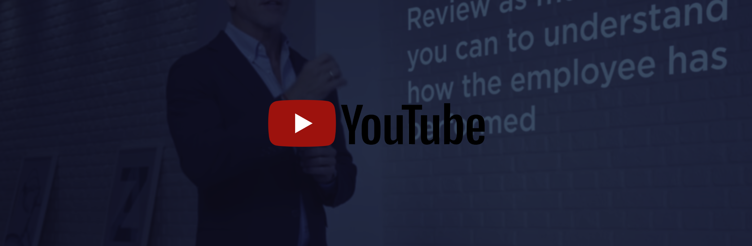 A woman in formal attire giving a presentation with a slide showing on the wall behind her, alongside the YouTube logo overlay.