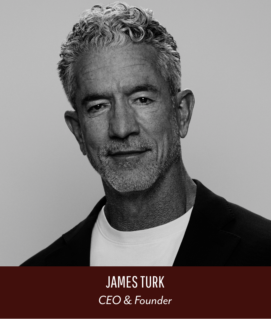 Black and white portrait of James Turk, middle-aged man with gray, curly hair, and a beard, dressed in a blazer and white shirt, smiling subtly at the camera.