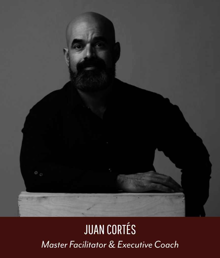 Black and white portrait of Juan Cortés, a man with a beard and bald head, wearing a dark shirt, posed resting his arm on a wooden block against a plain background. Text overlay reads 'Juan Cortés, Master Facilitator & Executive Coach.'