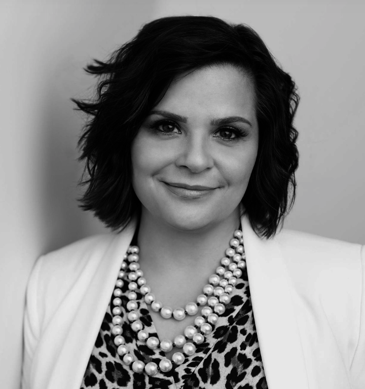 Black and white portrait of a woman with wavy, shoulder-length hair, wearing a pearl necklace, a leopard-print top, and a light-colored blazer, smiling softly at the camera.