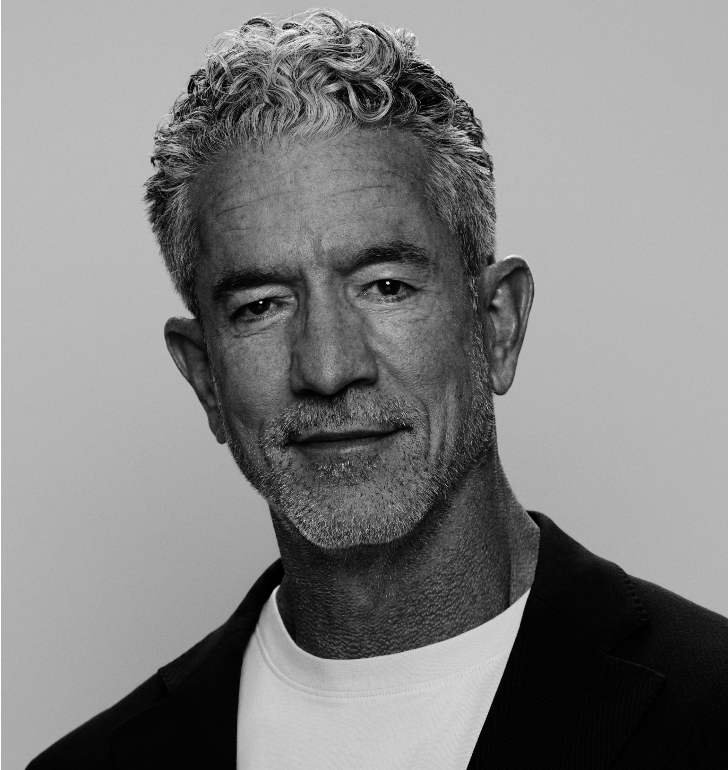 A black and white portrait of a middle-aged man with curly gray hair, trimmed beard, wearing a blazer and a white shirt.