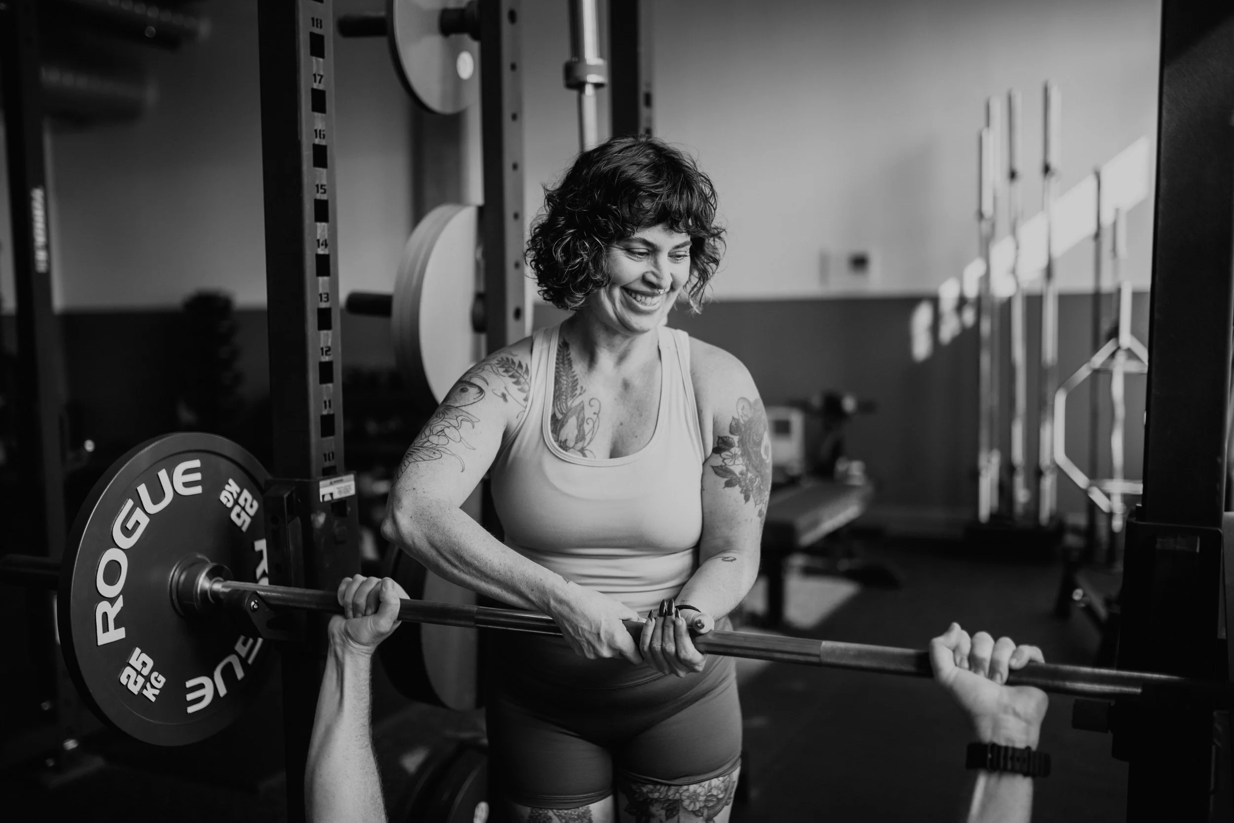A woman with curly hair lifting a barbell across her shoulders in a gym.