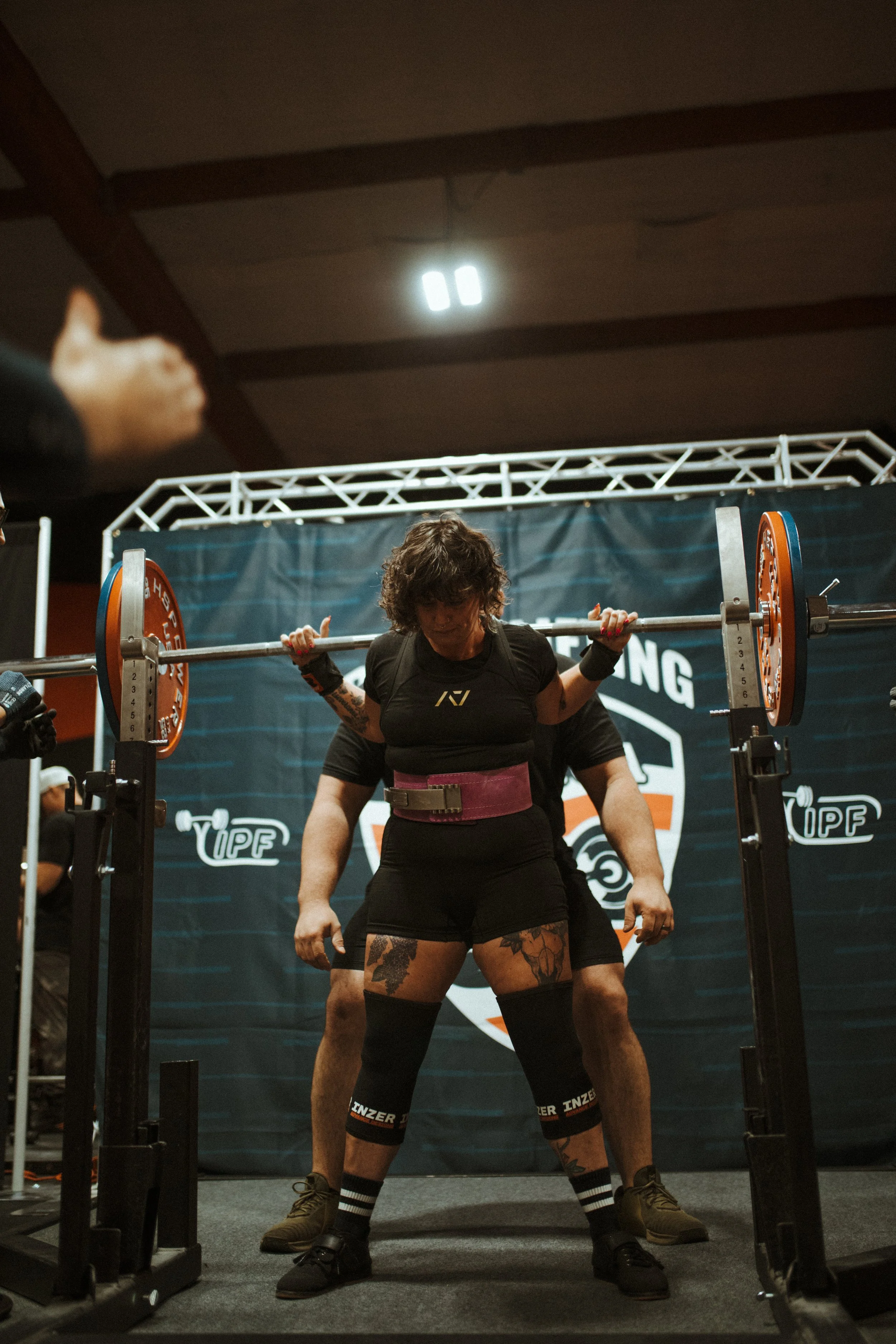 Female weightlifter in black attire with tattoos on thighs and resistance bands around knees, preparing to lift a barbell during competition.