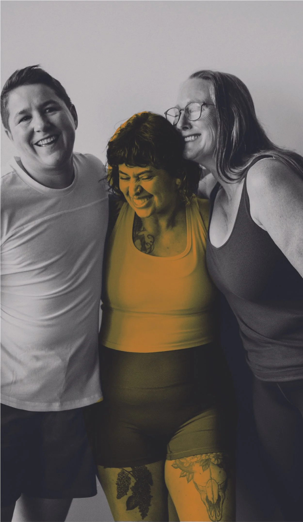 Three people smiling and hugging against a plain wall, with one person's tattoos and colored shirt standing out.