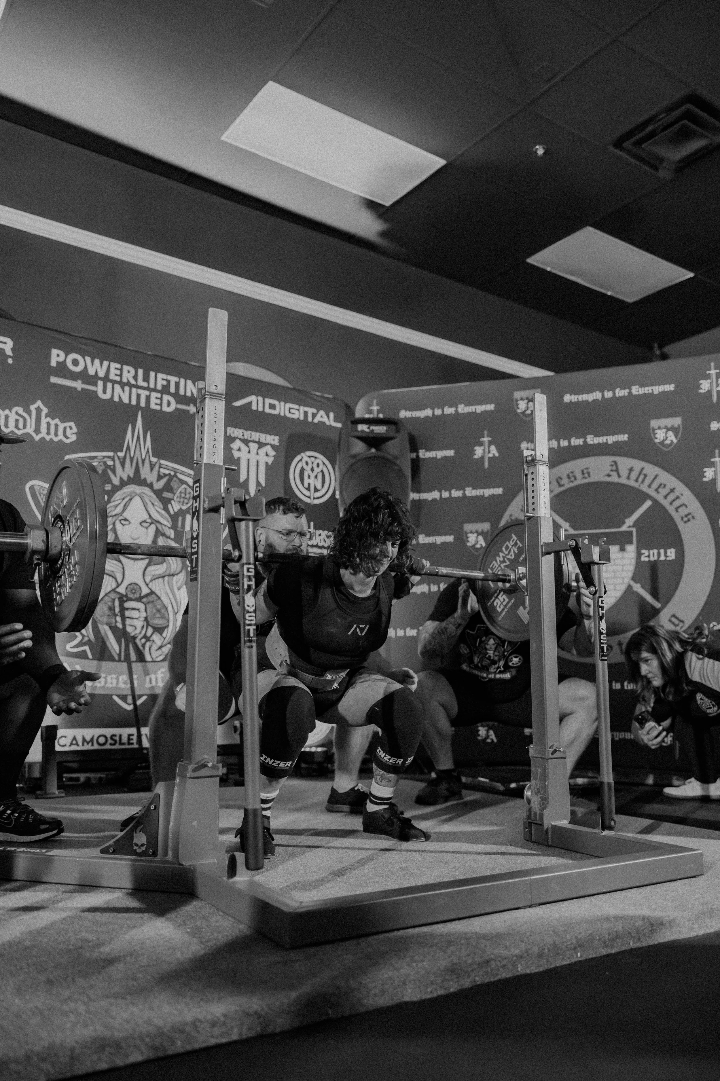 A woman participating in a powerlifting competition, squatting with a loaded barbell on her shoulders, surrounded by spotters and officials, with an intense expression showing effort.