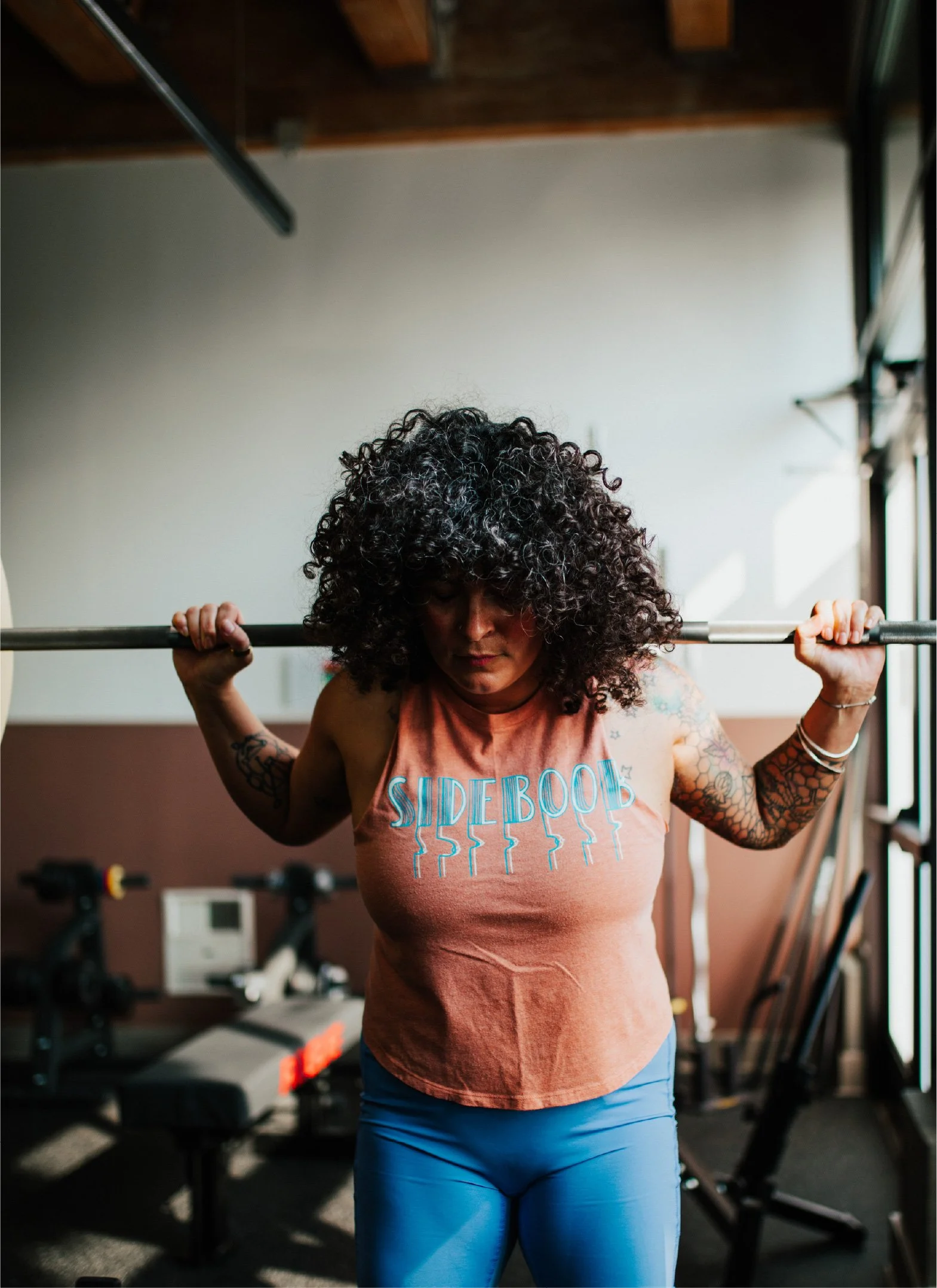 A woman with curly hair, wearing a sleeveless shirt and blue leggings, is lifting a barbell in a gym.