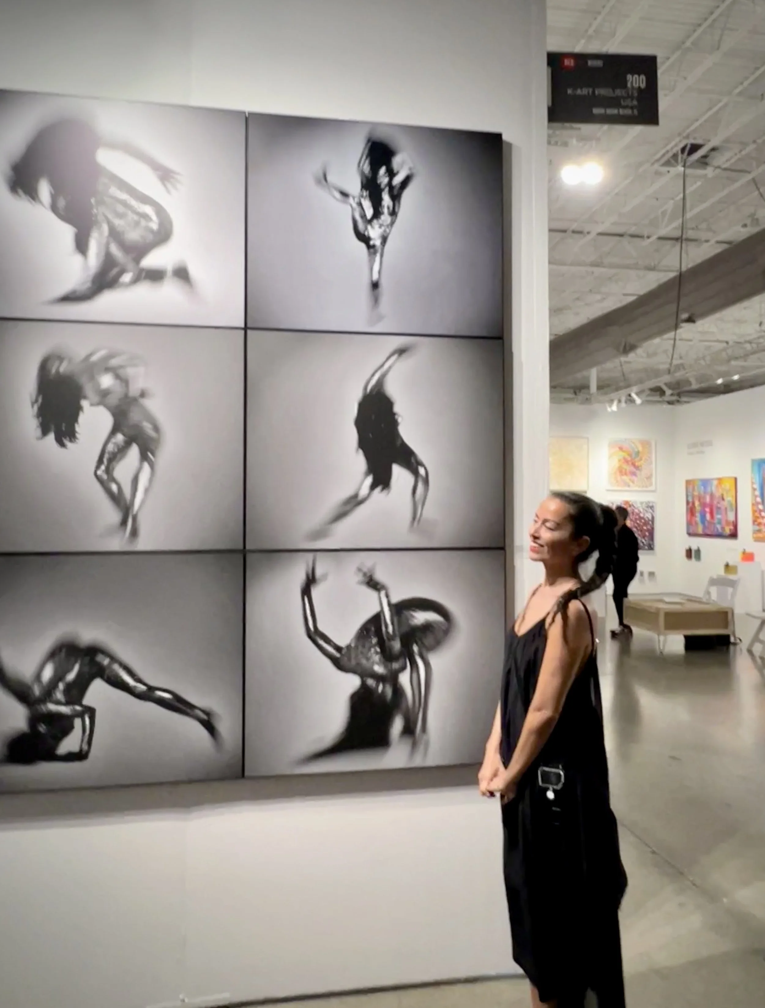photographer Nana Klimek Tinley standing in a gallery in front of six black-and-white photographic works capturing blurred dance movement, shown during Miami Art Week 2023.