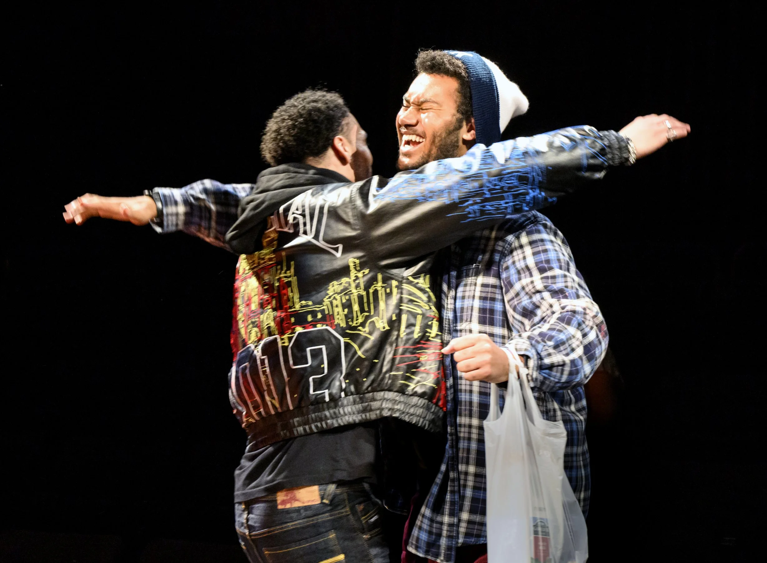 Two men hugging and smiling on a black background, one holding a plastic bag.