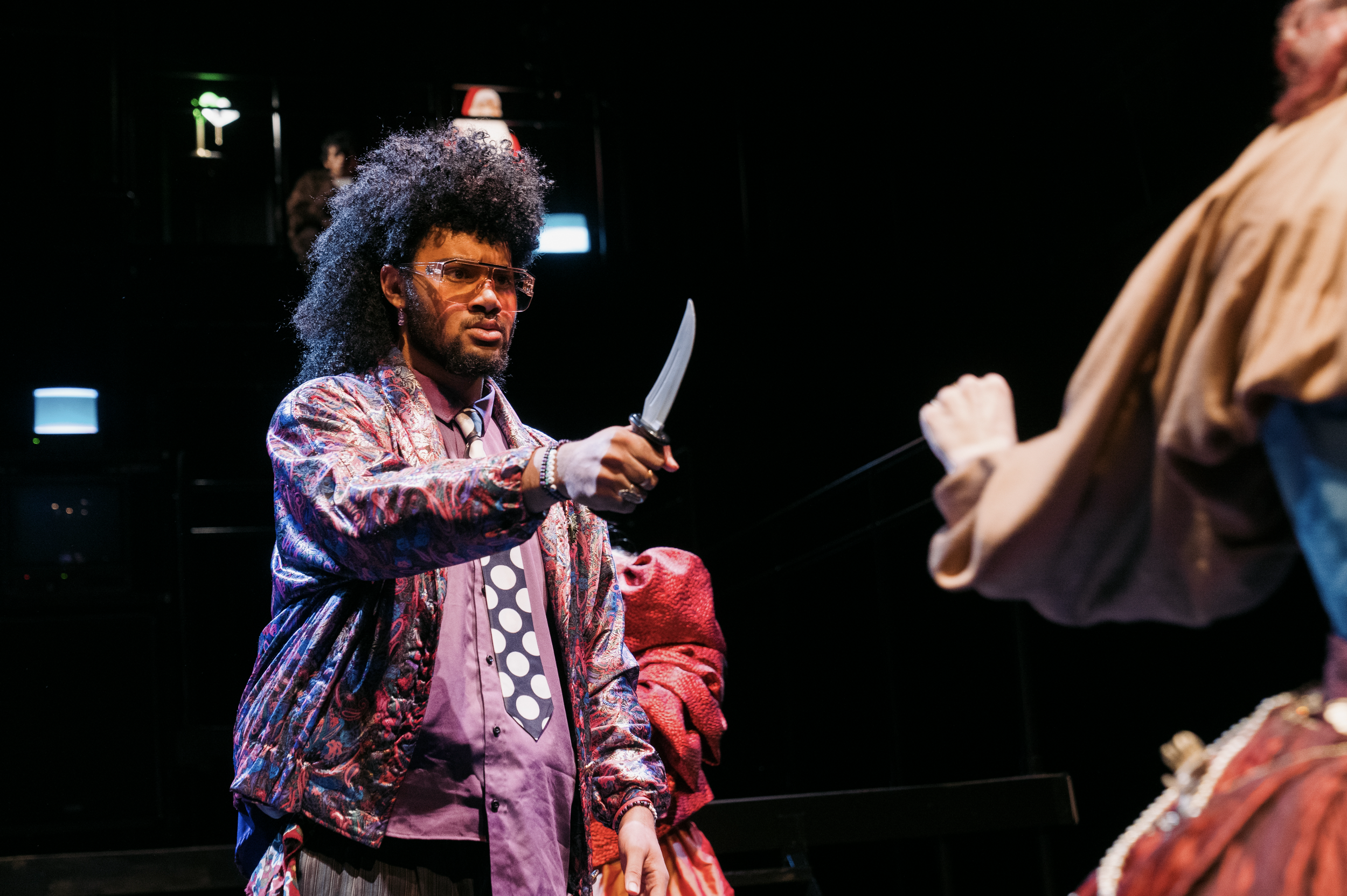 A man with an afro hairstyle and glasses is holding a large knife, pointing it towards another person's chest during a theatrical performance.