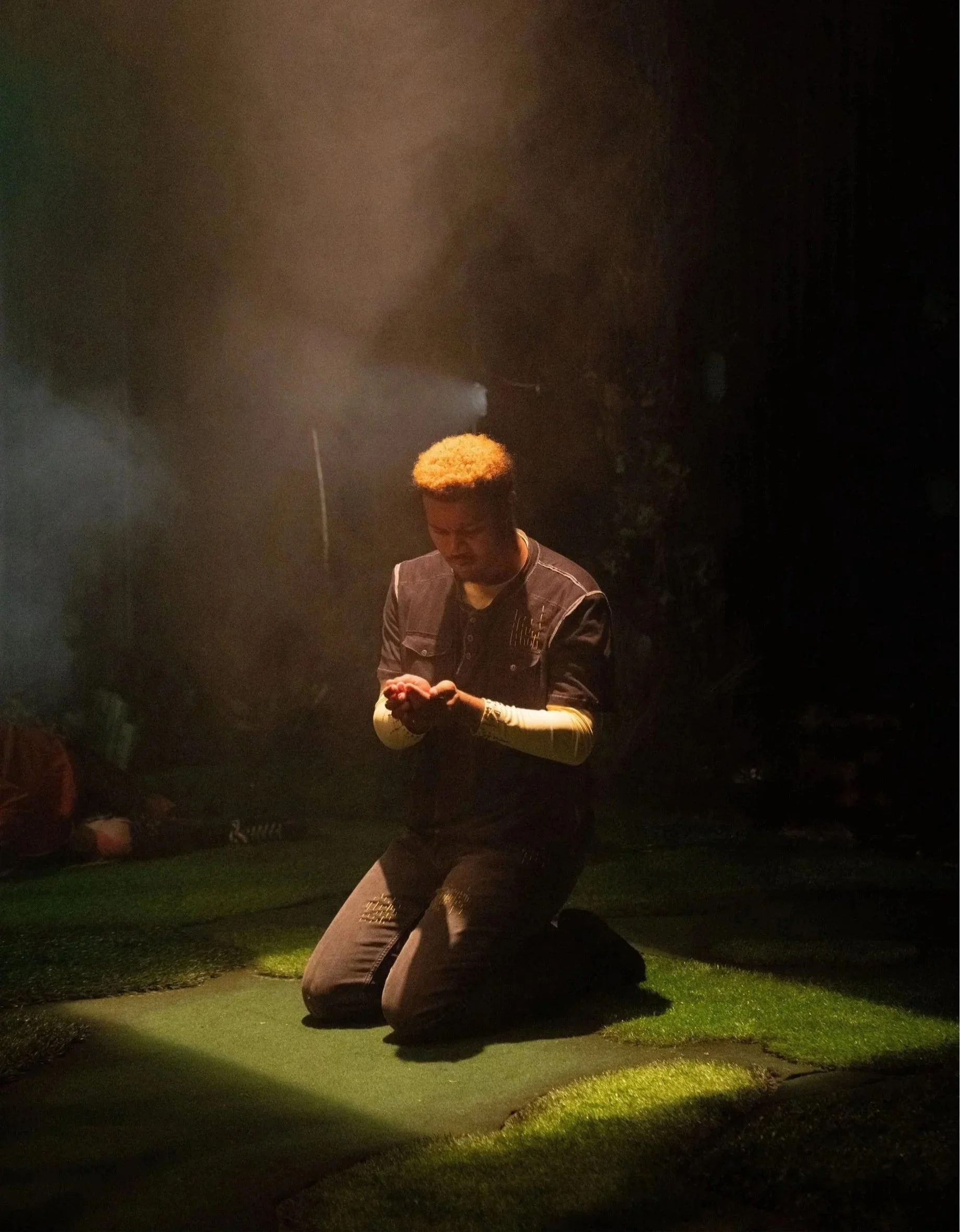 A person with blonde hair kneeling on the ground with hands together, illuminated by a spotlight in a dark setting.