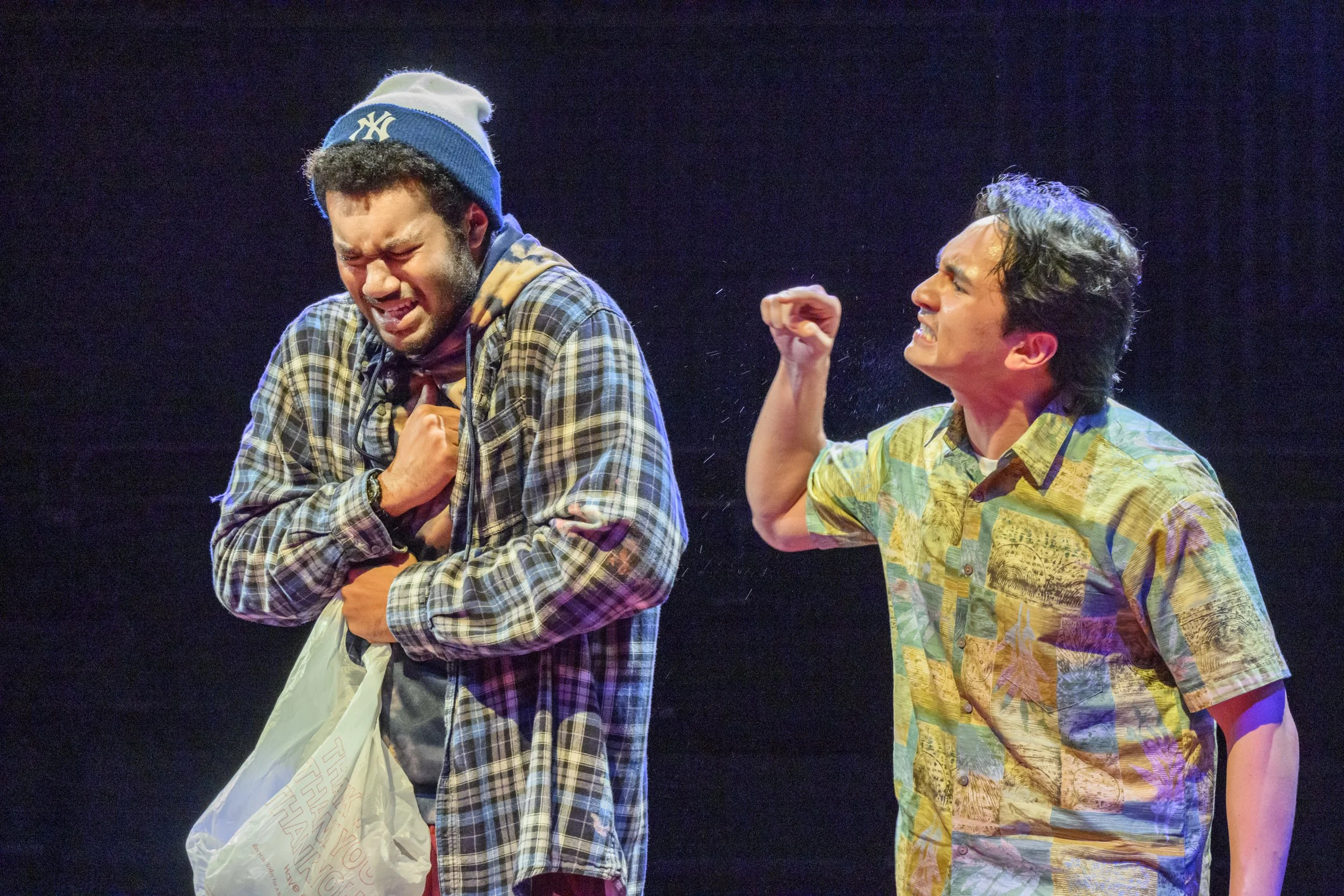 Two men are on stage, appearing emotional. The man on the left is wearing a plaid shirt, a Seattle Seahawks beanie, and holds a plastic bag, clutching his chest with a distressed expression. The man on the right is wearing a colorful short-sleeve shirt, and is gesturing passionately with his clenched fist raised, looking intensely at the other man.