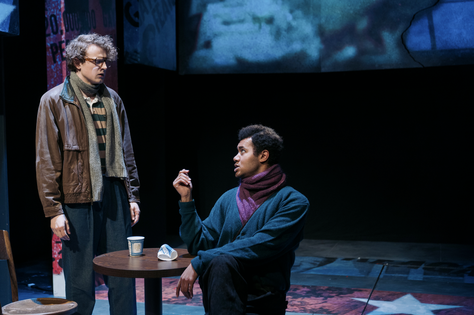 Two men engaged in a serious conversation on a stage. One is standing, wearing glasses, a leather jacket, and a striped shirt, while the other is sitting, gesturing with his hand, wearing a teal sweater and a purple scarf. There is a small round table with a paper cup, a face mask, and some items on it, with a dark stage background and a screen displaying abstract visuals above them.