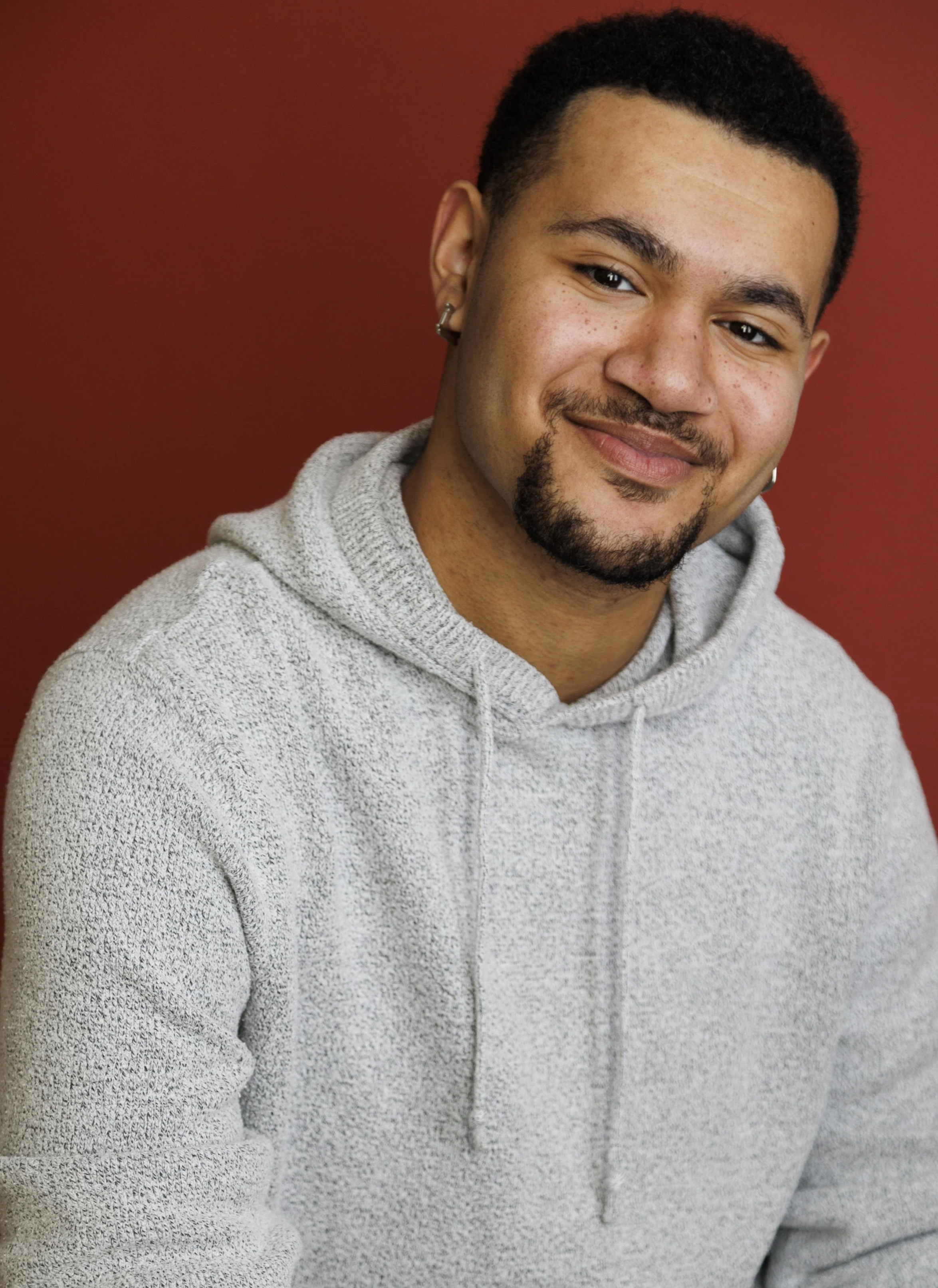Portrait of a smiling young man with light brown skin, short dark hair, a goatee, wearing earrings, and a light gray hoodie, against a red background.