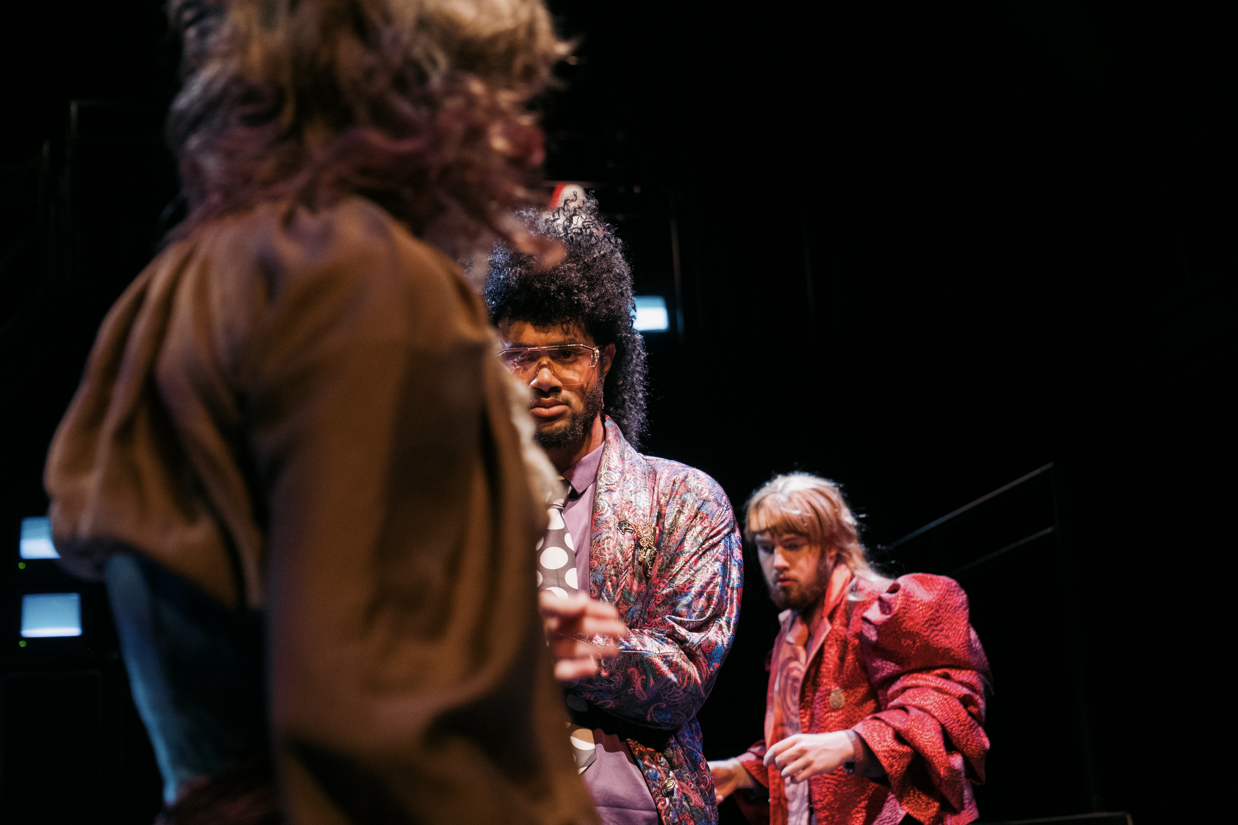 Three individuals, two men and one woman, are engaged in conversation or observation in a dimly lit environment. The man in the foreground has curly black hair, a beard, and is wearing glasses, a patterned jacket, and a shirt. The woman in the background has blonde hair and is dressed in a red textured jacket or coat. The other woman has her back to the camera with brown, wavy hair and is wearing a brown jacket.