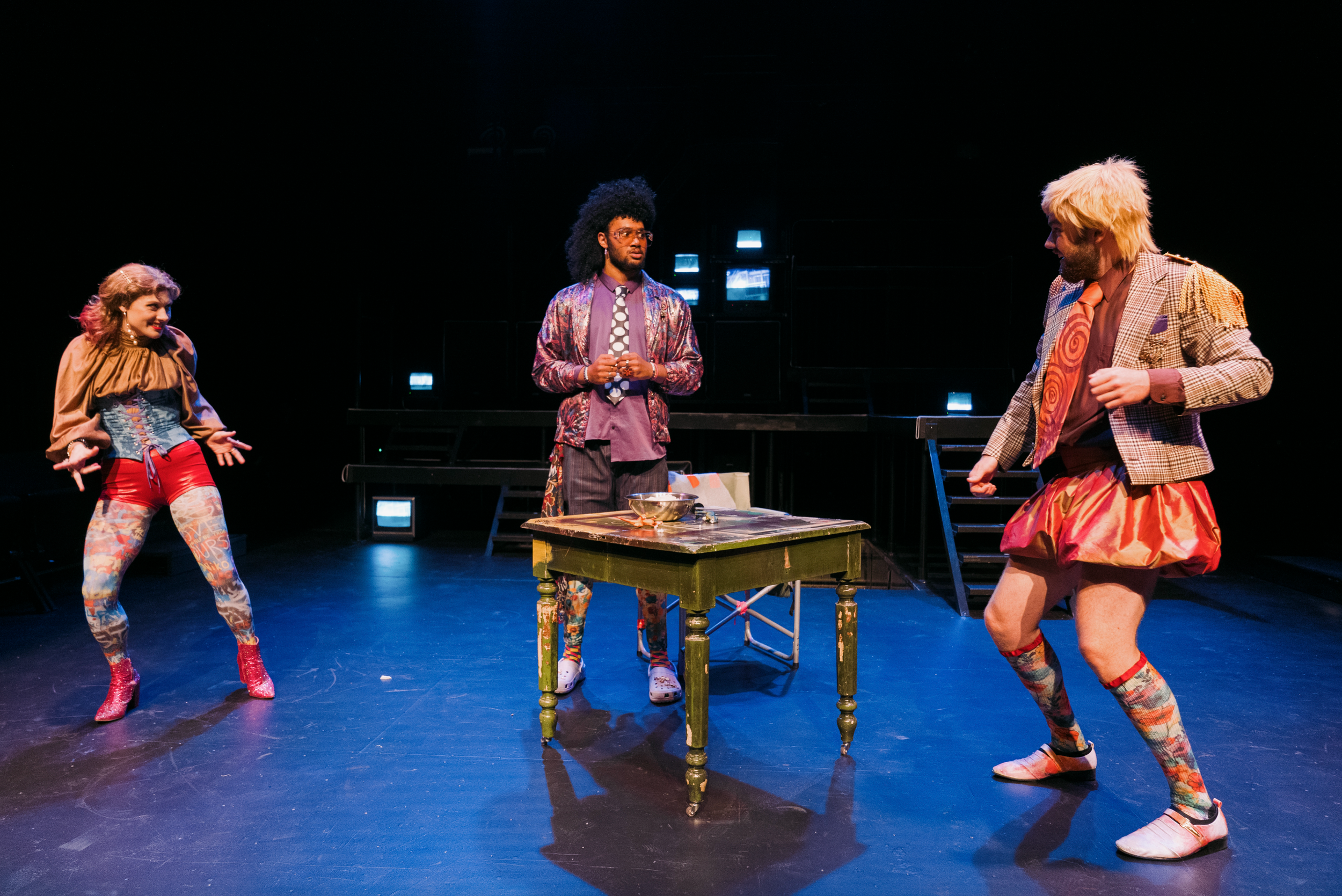 Three performers in colorful costumes on stage during a theatrical production, with a small table and bright stage lights.